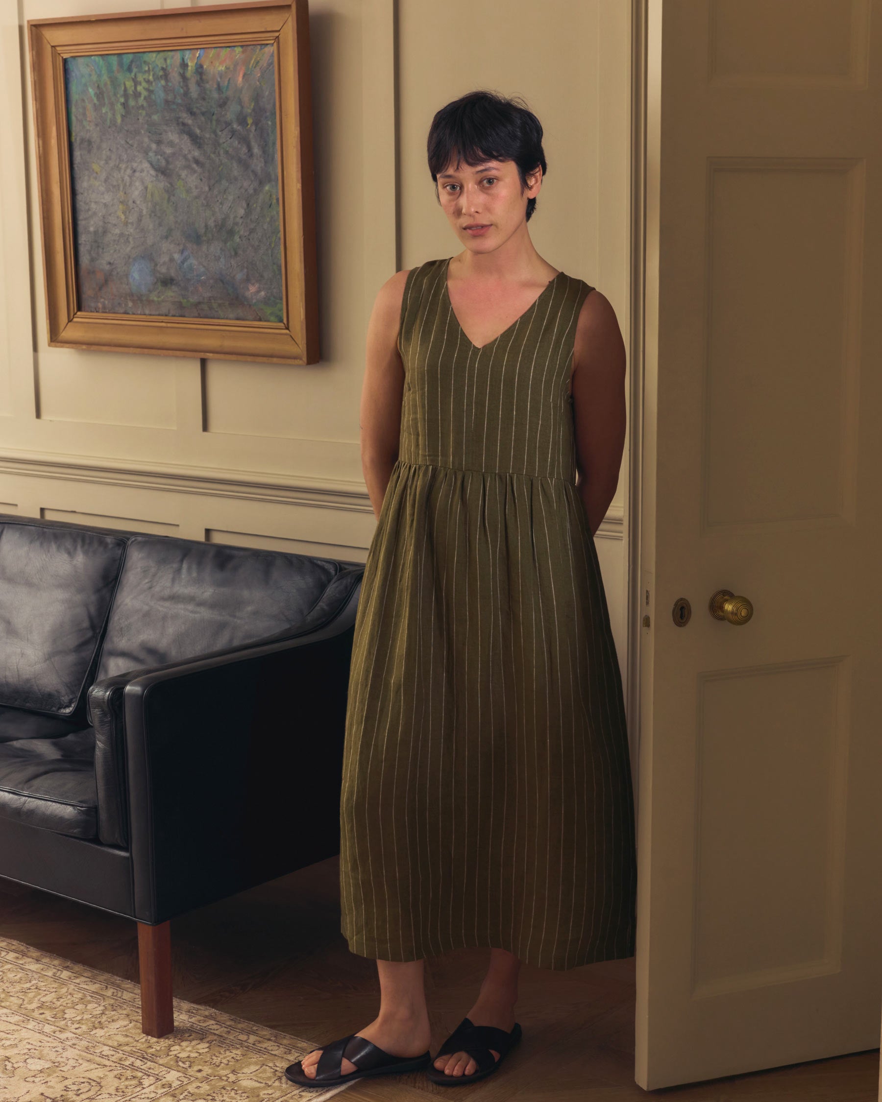 A woman with short dark hair stands indoors by an open door, wearing the Pyne and Smith Clothiers No.21 V Neck Sleeveless Linen Dress in Kelp Green Stripe with black sandals. A dark sofa and framed painting are behind her.