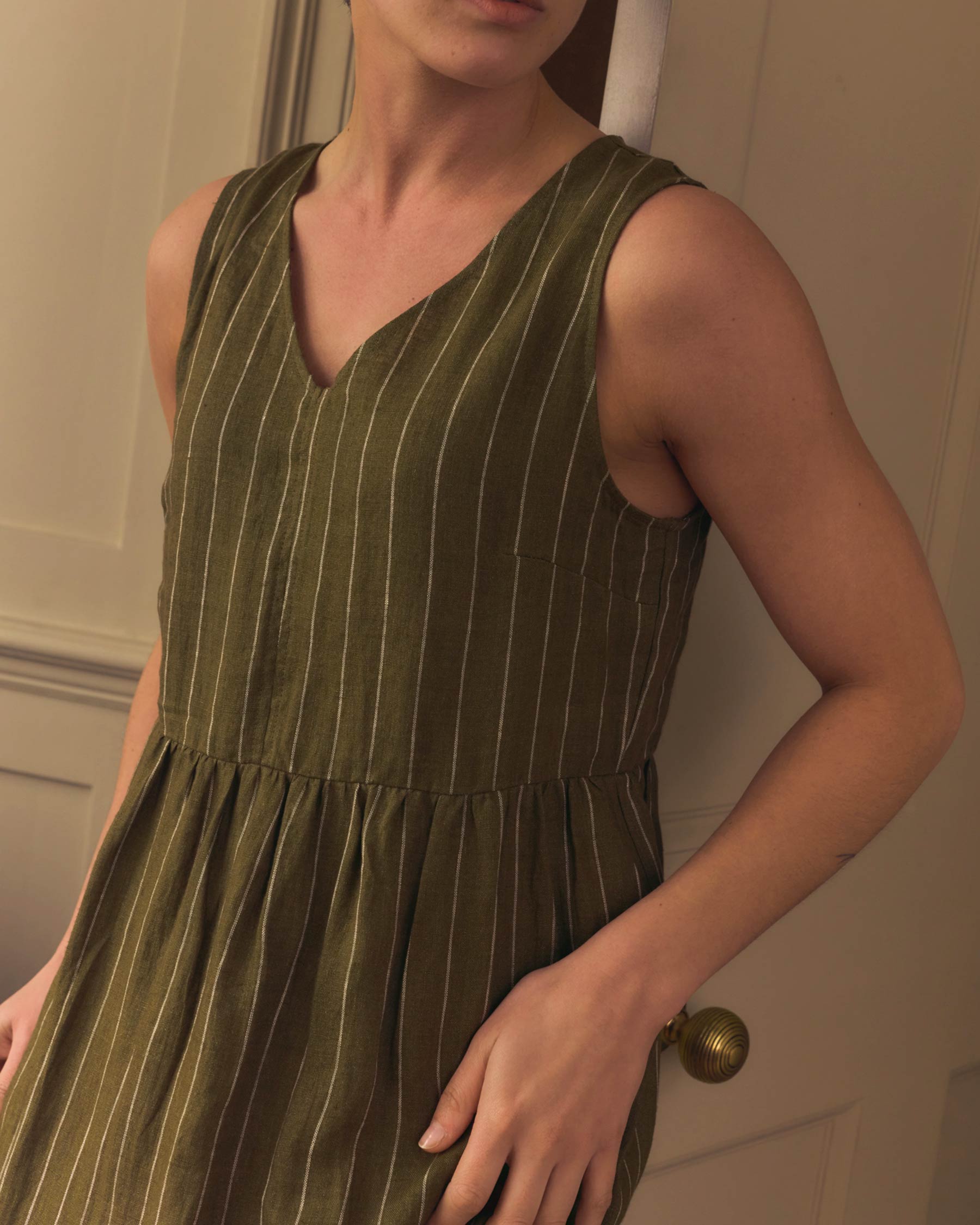 A person, visible from the shoulders down, leans against a cream-colored door wearing the Pyne and Smith Clothiers No.21 V Neck Sleeveless Linen Dress in Kelp Green Stripe with white vertical stripes.
