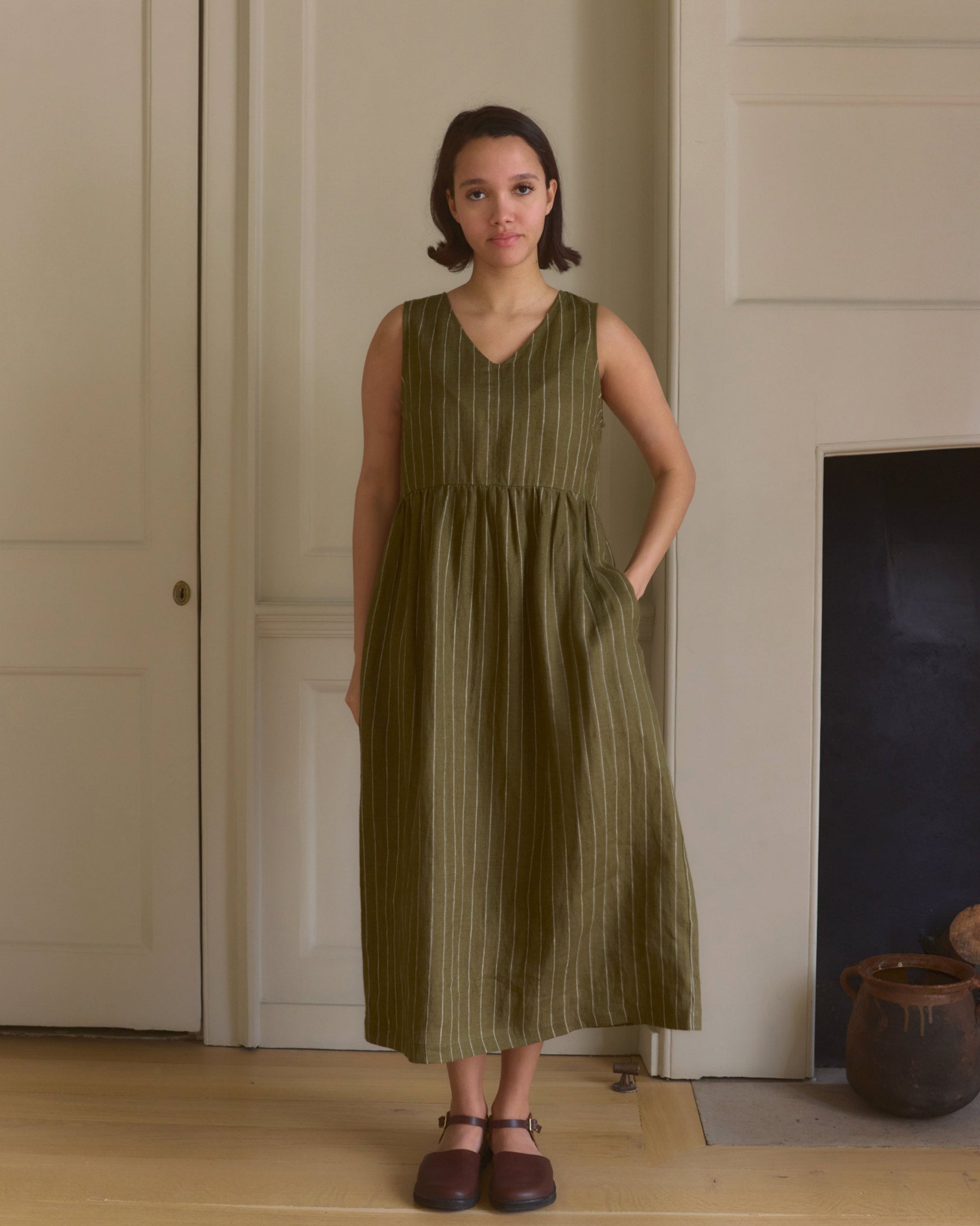 A woman stands indoors wearing the Pyne and Smith Clothiers No.21 V Neck Sleeveless Linen Dress in Kelp Green Stripe, paired with brown shoes. She has short hair, one hand in her pocket, against a neutral wall and wooden floor.
