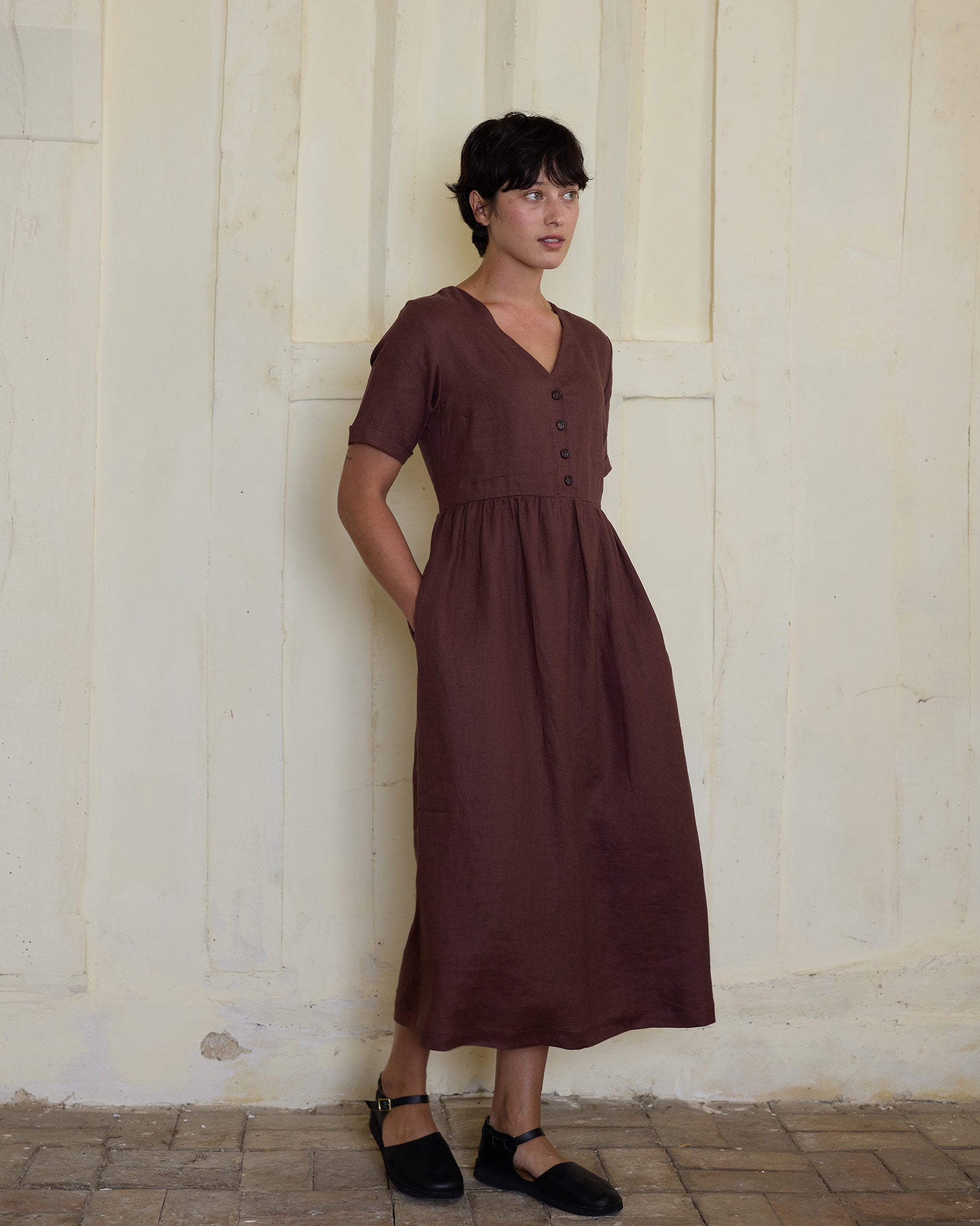 A woman with short dark hair models the Pyne and Smith Clothiers No.31 V Neck, Full Length Linen Dress in Marron, styled with black strap shoes against a light-toned textured wall—an effortless embrace of sustainable fashion.