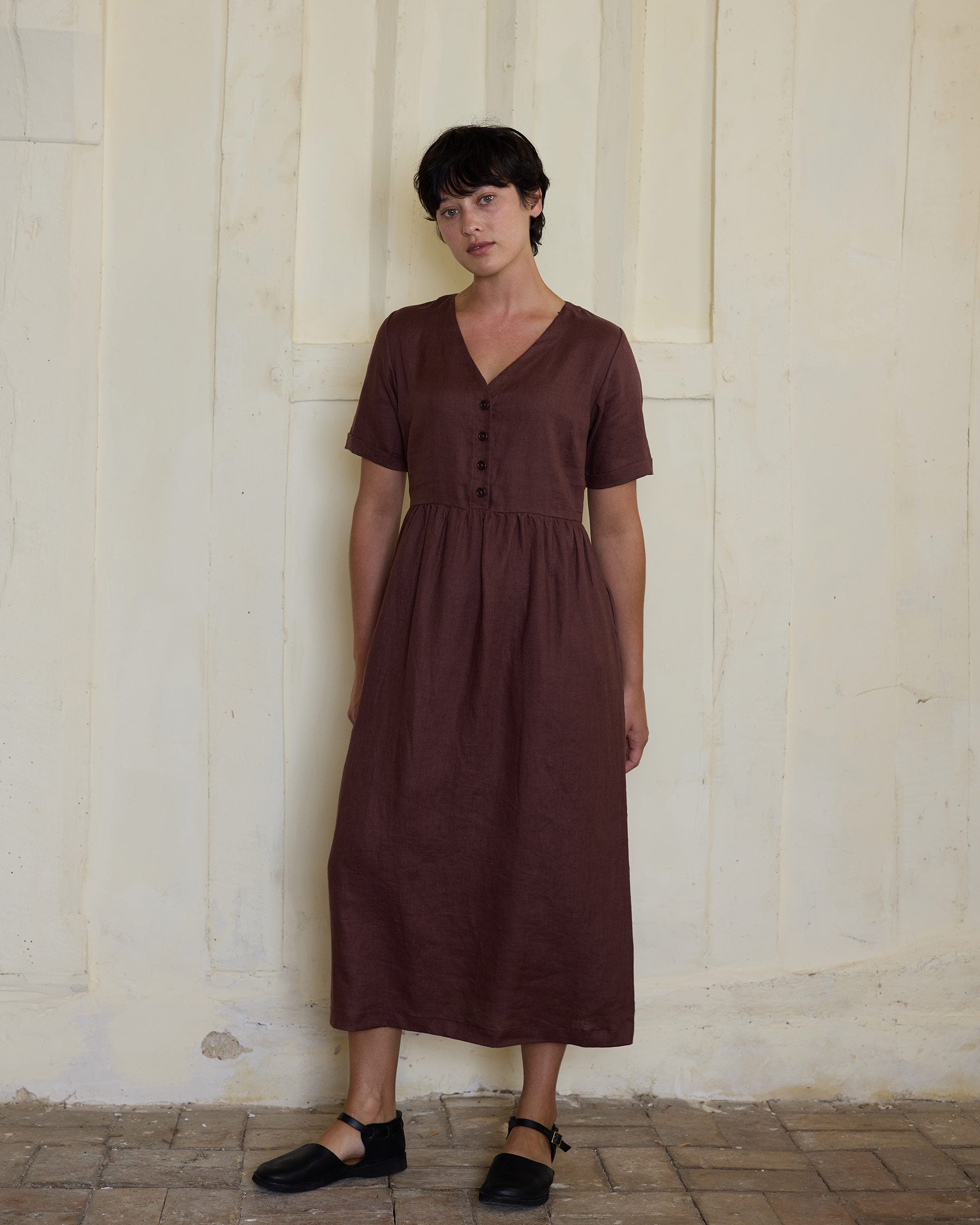A person with short, dark hair wears the Pyne and Smith Clothiers No.31 V Neck, Full Length Linen Dress in Marron and black shoes, standing against a light-colored textured wall and tiled floor.