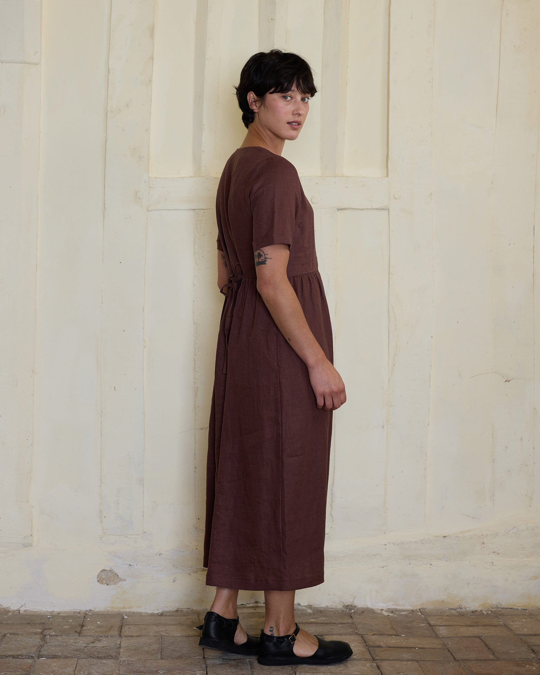 A person with short dark hair wears the Pyne and Smith Clothiers No.31 V Neck Full Length Linen Dress in Marron and black shoes, standing sideways on a tiled floor against a cream textured wall, embodying sustainable fashion.