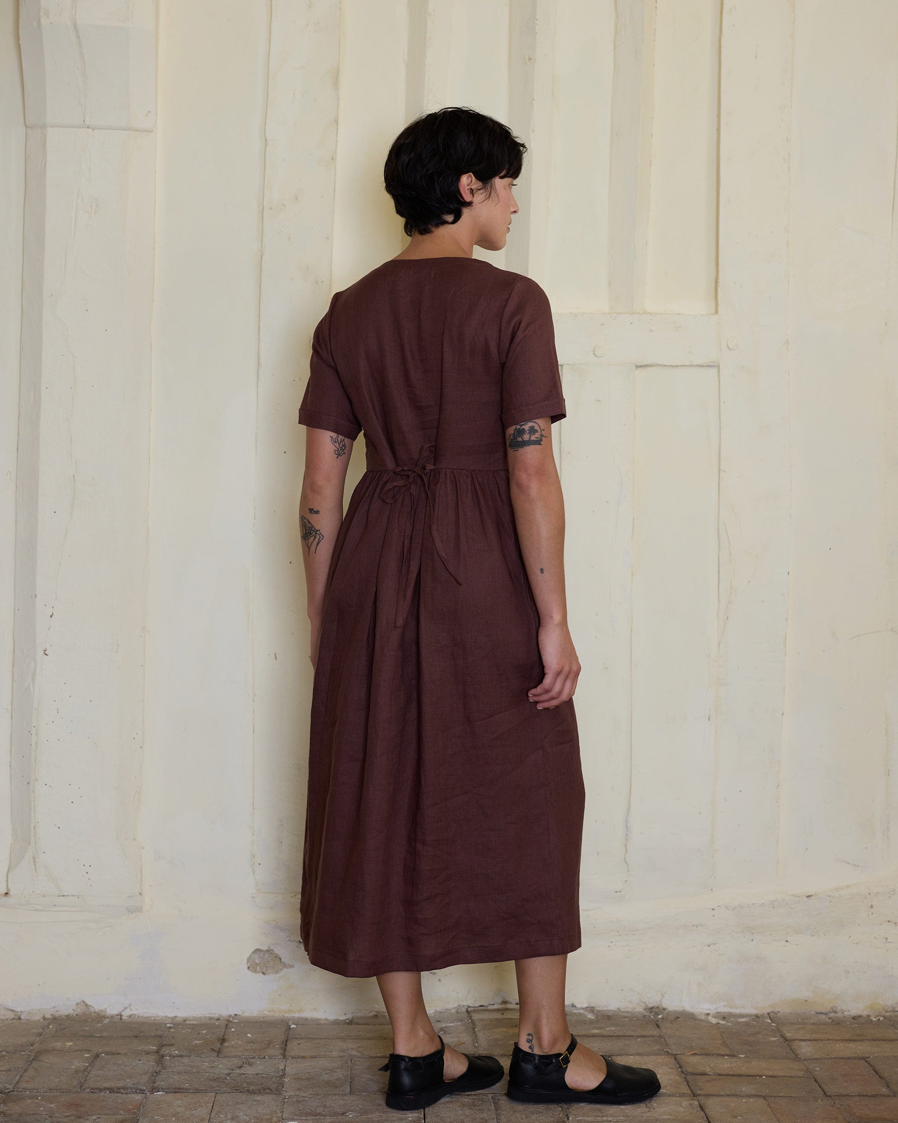 A person with short dark hair faces away indoors, wearing the Pyne and Smith Clothiers No.31 V Neck, Full Length Linen Dress in Marron. The dress features short sleeves, back tie, and ankle-length hem. Light textured wall and tiled floor background.