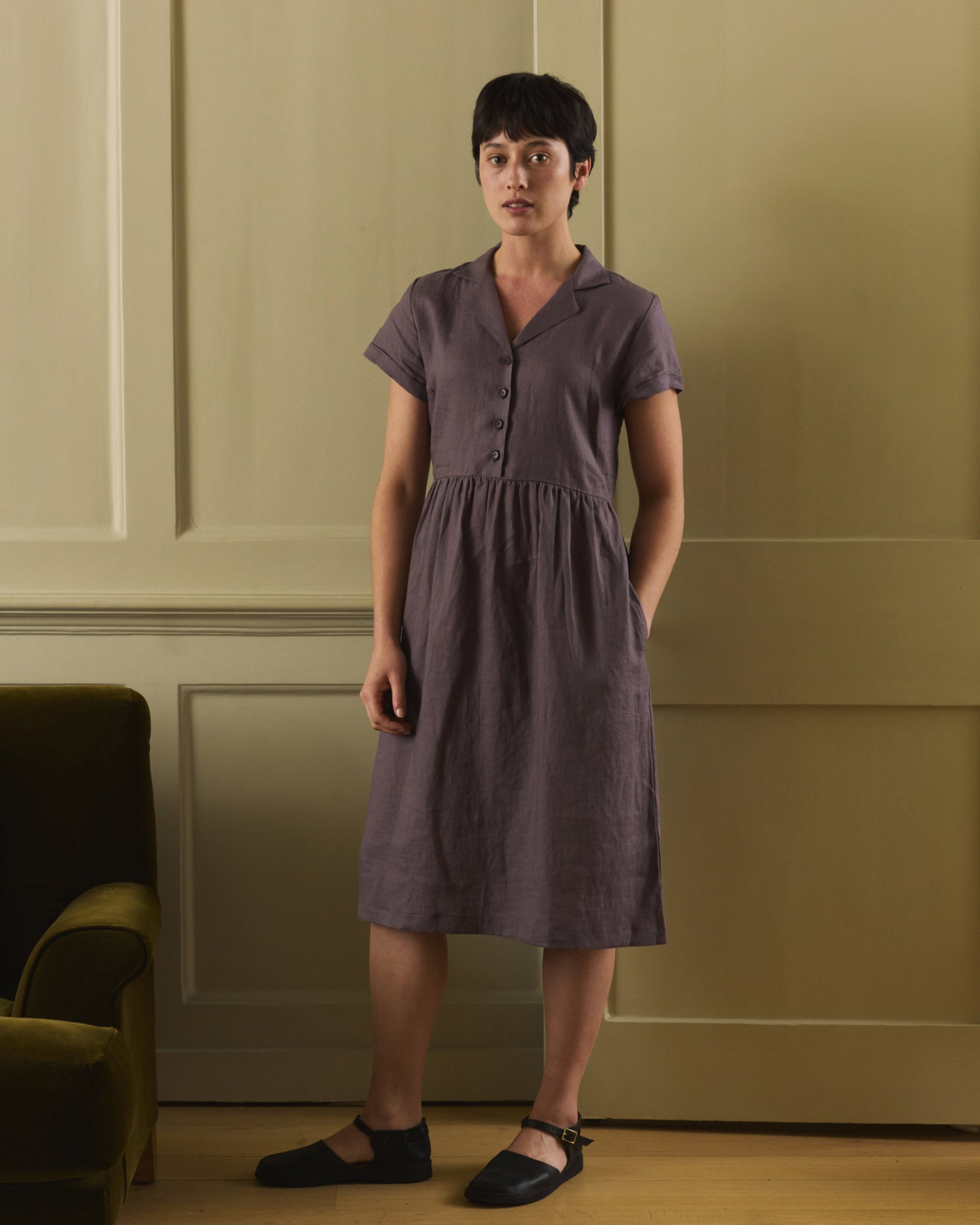A woman with short dark hair stands indoors against a pale wall, wearing the No.30 V-neck, Collared Linen Dress in Plum Truffle by Pyne and Smith Clothiers with Corozo nut buttons and black flats, her hands in her pockets. An olive green chair is nearby.