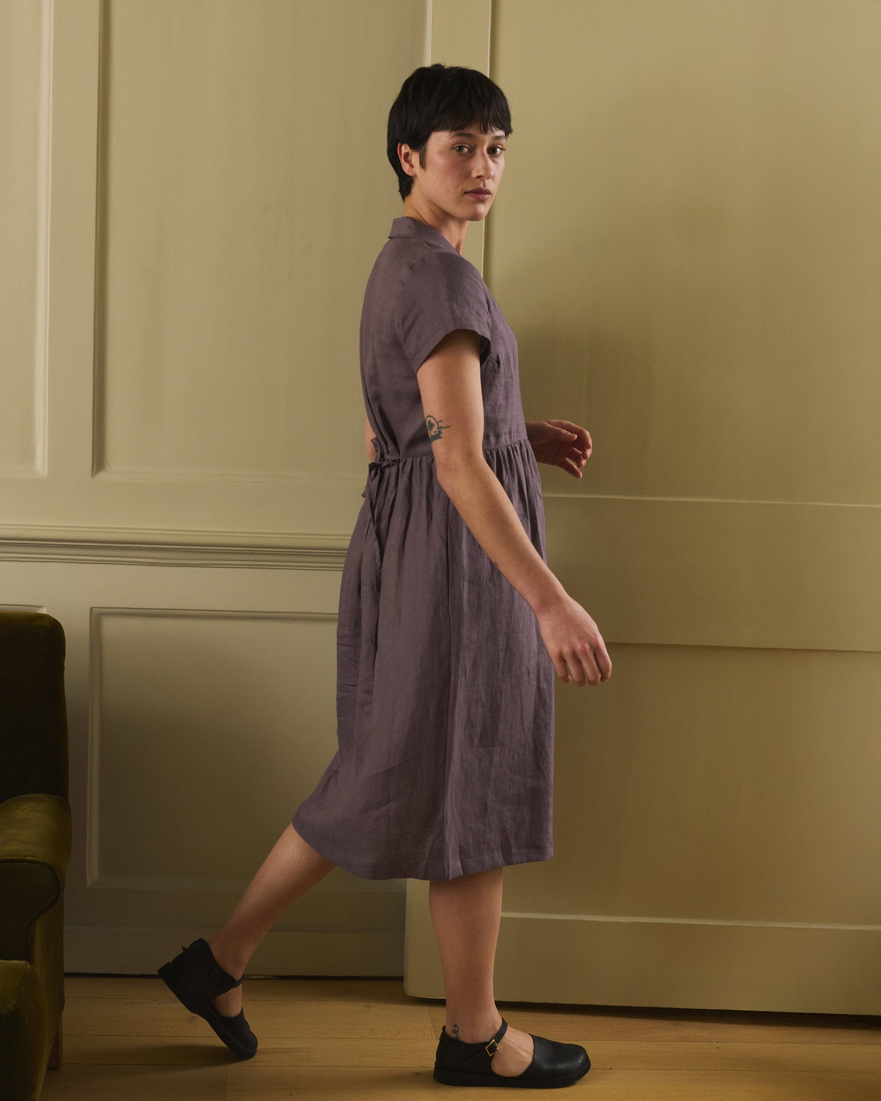 A person with short dark hair stands sideways, looking over their shoulder, wearing Pyne and Smith Clothiers No.30 V-neck, Collared Linen Dress in Plum Truffle in a softly lit room with beige paneled walls and a green sofa.