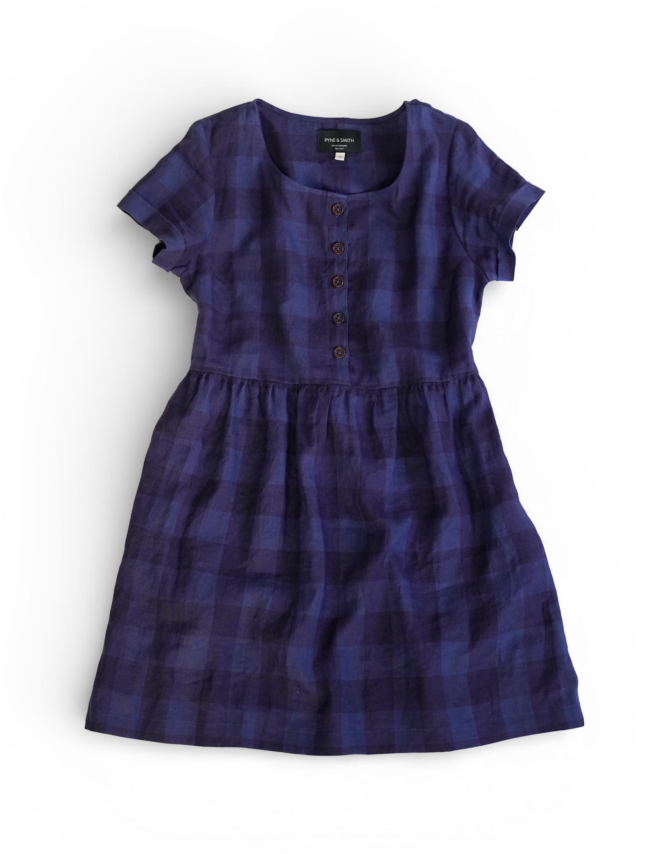 The No.10 Button Up Linen Dress in Midsommar Night Check by Pyne and Smith Clothiers features short sleeves, a fitted waist, knee-length cut, navy and black plaid pattern, and front buttons. It’s shown laid flat on a white background.