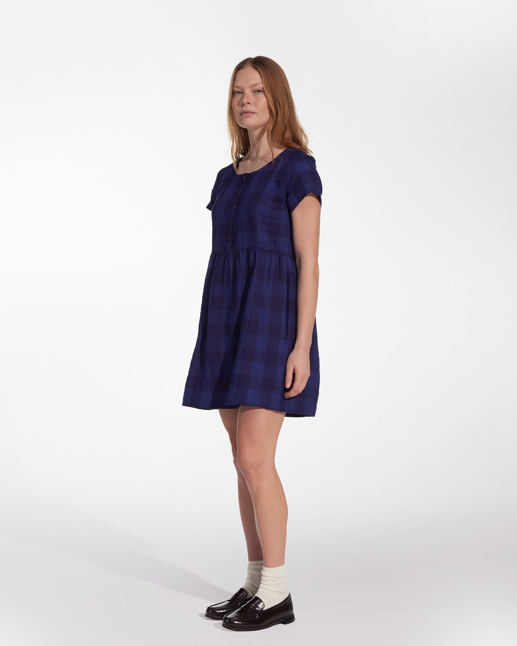 A person with long hair stands against a white background, wearing the Pyne and Smith Clothiers No.10 Button Up Linen Dress in Midsommar Night Check, paired with white socks and black loafers.