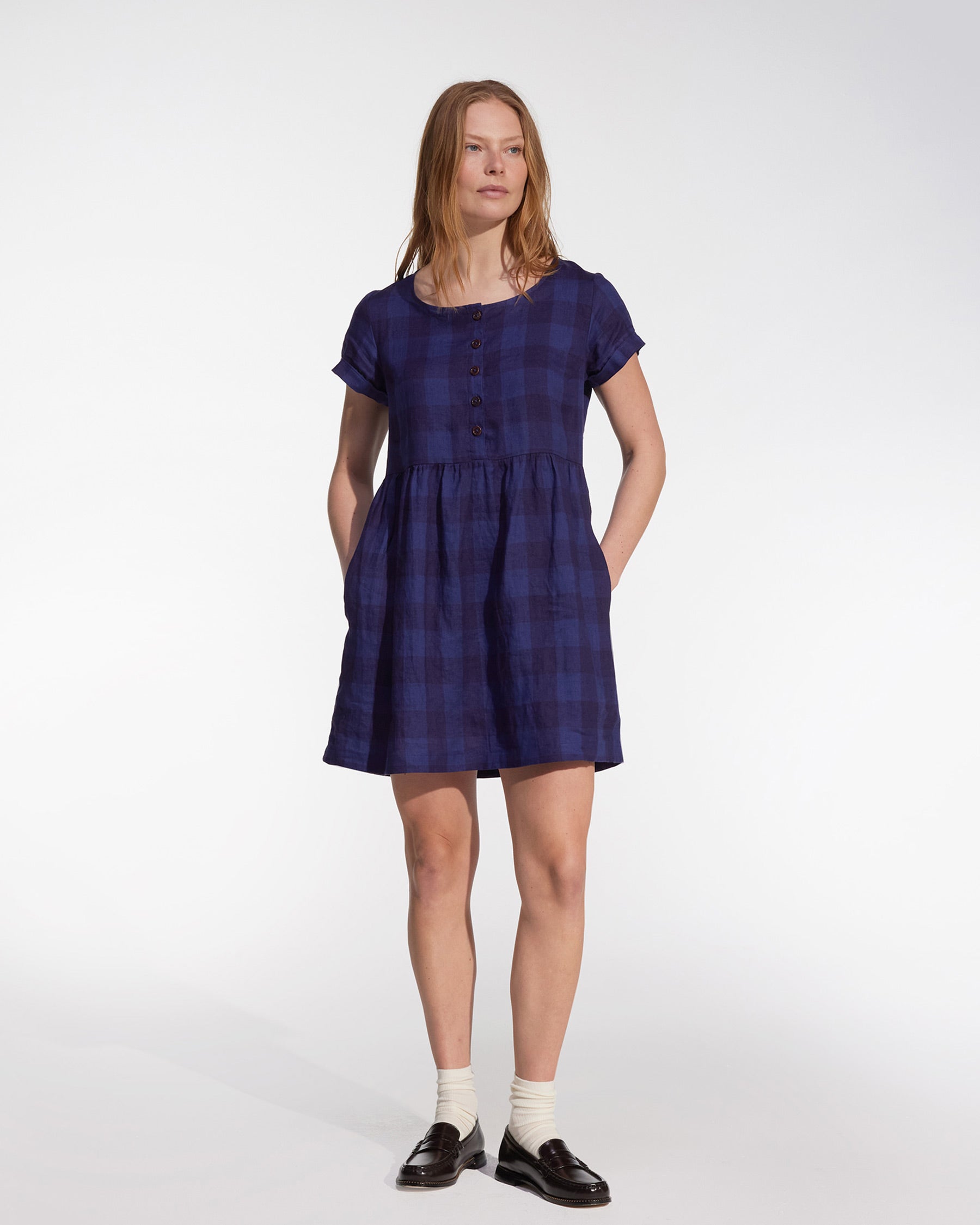 A woman stands with hands in her pockets, wearing the No.10 Button Up Linen Dress in Midsommar Night Check by Pyne and Smith Clothiers, paired with white ankle socks and black loafers against a plain white background.