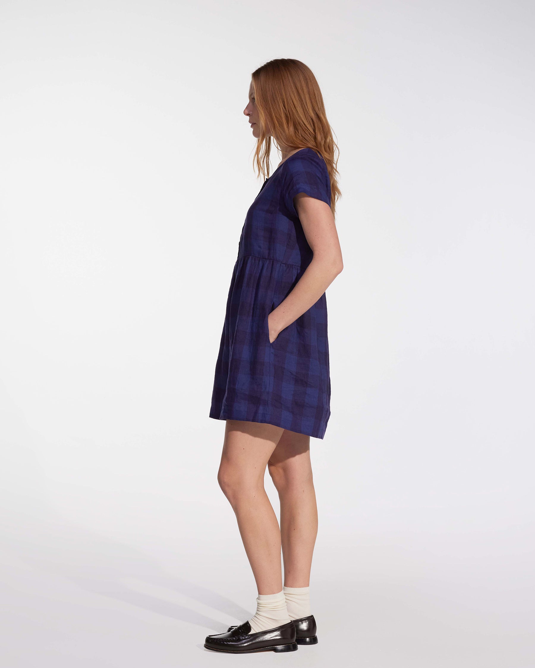 A woman stands in profile against a white background, wearing the Pyne and Smith Clothiers No.10 Button Up Linen Dress in Midsommar Night Check with pockets, white socks, black loafers, and loose hair. Her hands are tucked into her pockets.