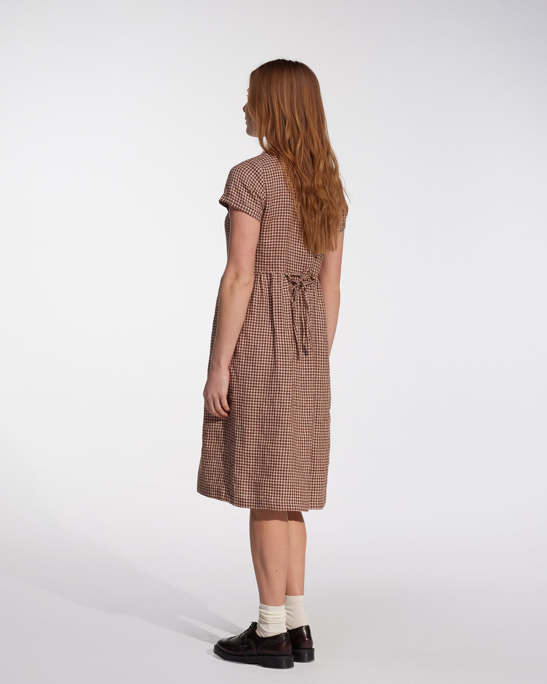 A woman with long, straight, reddish-brown hair stands facing away in the Pyne and Smith Clothiers No.11 Classic Button Up Linen Dress in Topsham Check, featuring short sleeves, a knee-length hem, a back tie, white socks, and dark shoes against a plain light background.