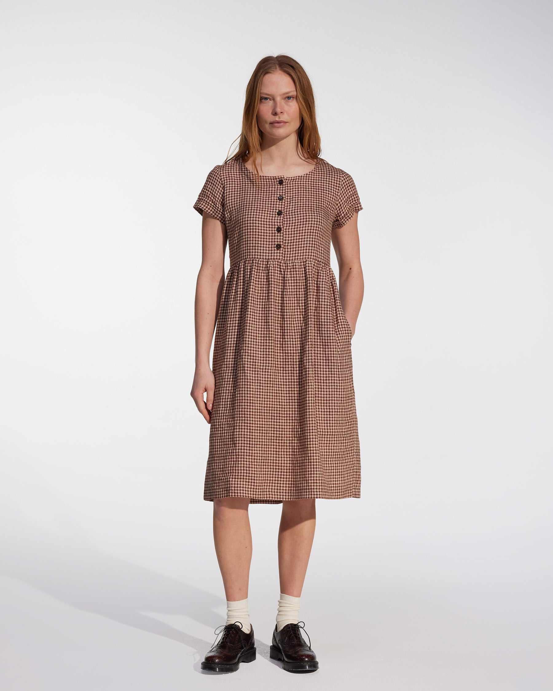 A woman stands against a white background wearing the Pyne and Smith Clothiers No.11 Classic Button Up Linen Dress in Topsham Check, paired with white socks and black shoes.