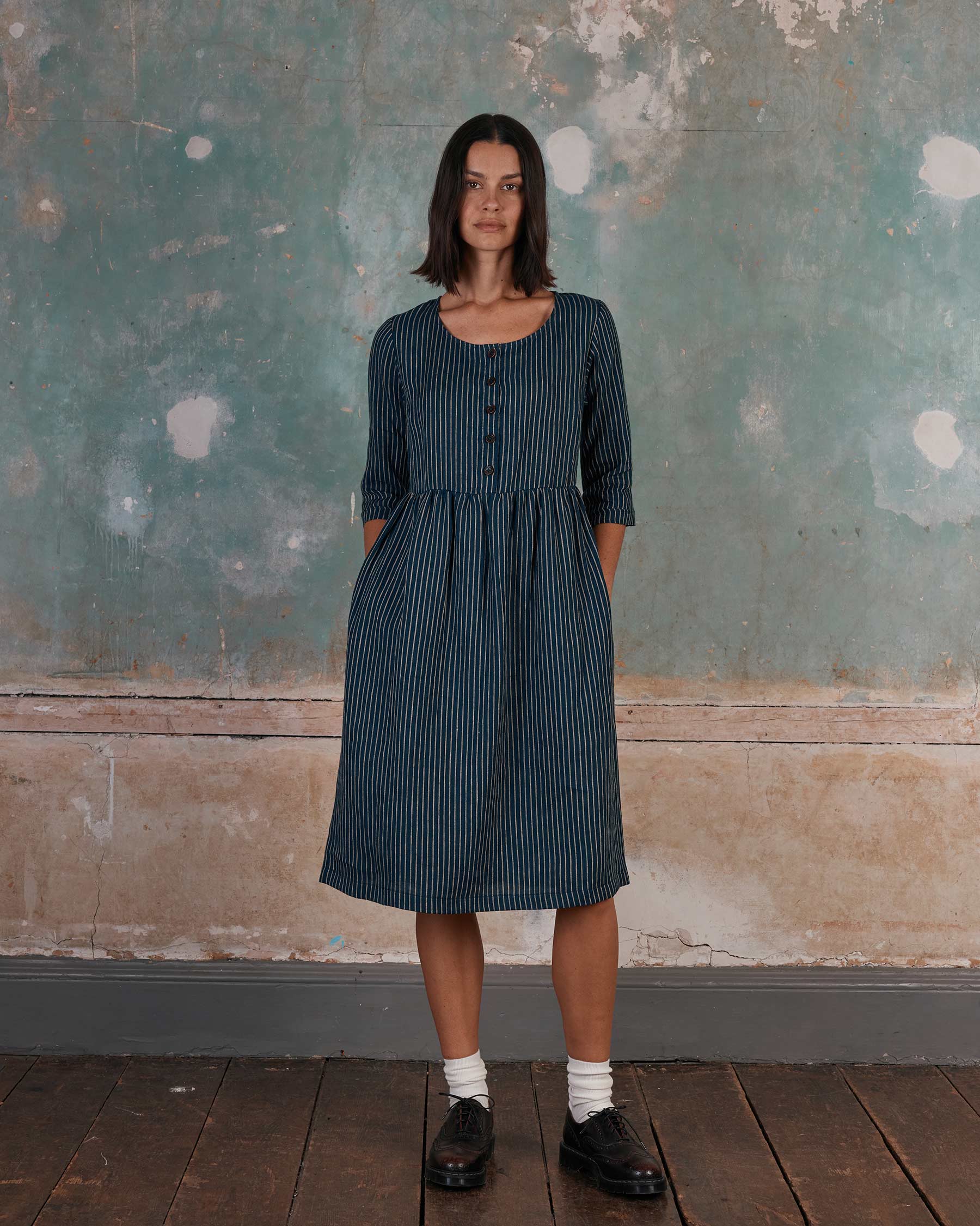 A woman in the Pyne and Smith Clothiers No.12 Button Up, Linen Midi Dress in Mariner Stripe stands by a weathered wall. Made in California, this medium-weight linen dress features pockets. She pairs it with white socks and black shoes.