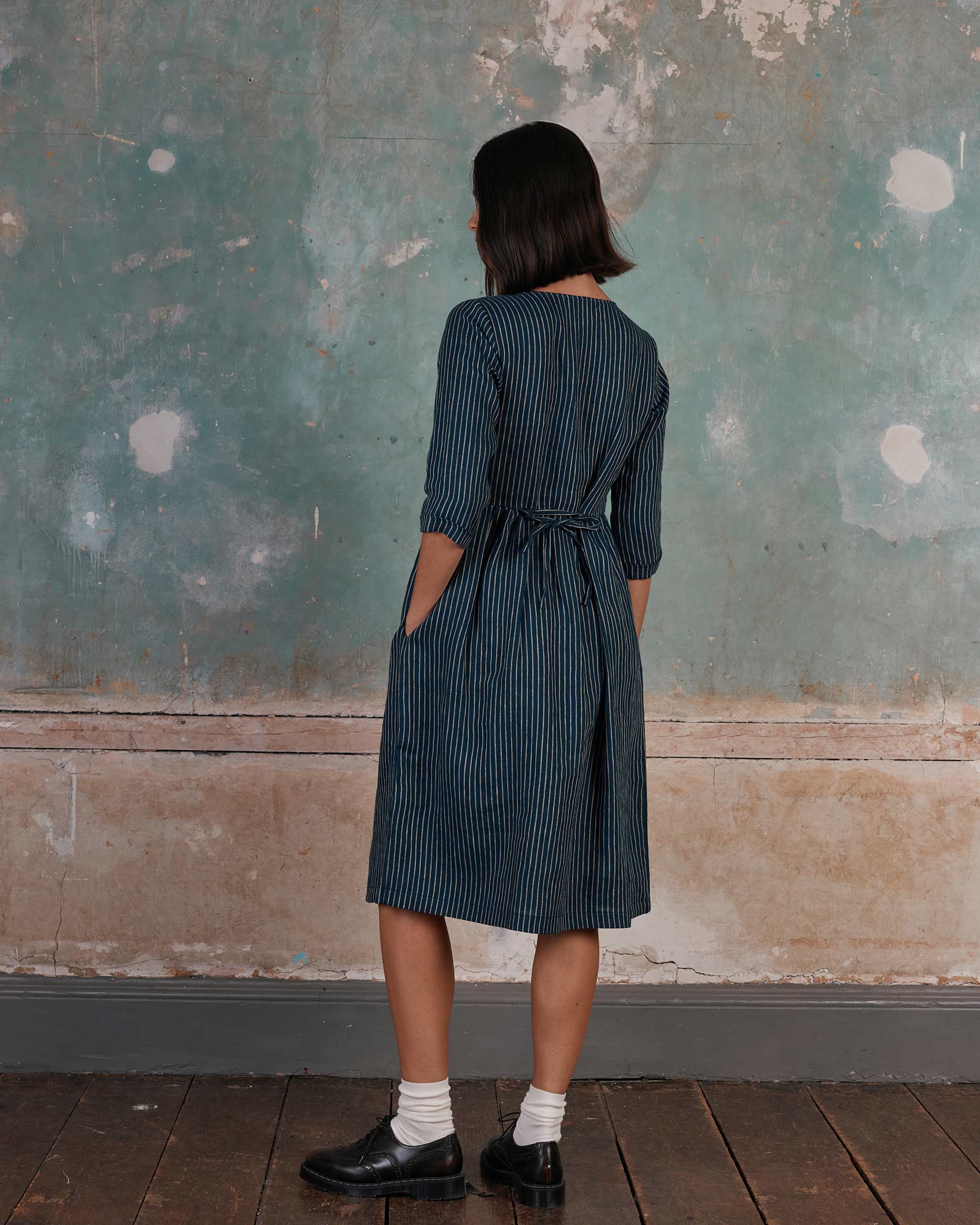 A woman faces away from the camera, wearing the Pyne and Smith Clothiers No.12 Button Up Linen Midi Dress in Mariner Stripe. The belted, three-quarter sleeve dress pairs with black shoes against a textured wall. Made in California.