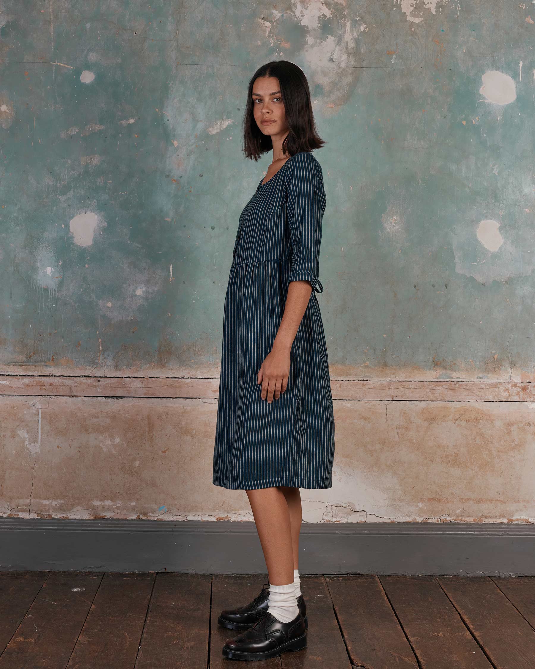 A woman with shoulder-length dark hair stands sideways against a distressed teal and beige wall, wearing the No.12 Button Up, Linen Midi Dress in Mariner Stripe by Pyne and Smith Clothiers, with white socks and black shoes on a dark wooden floor.
