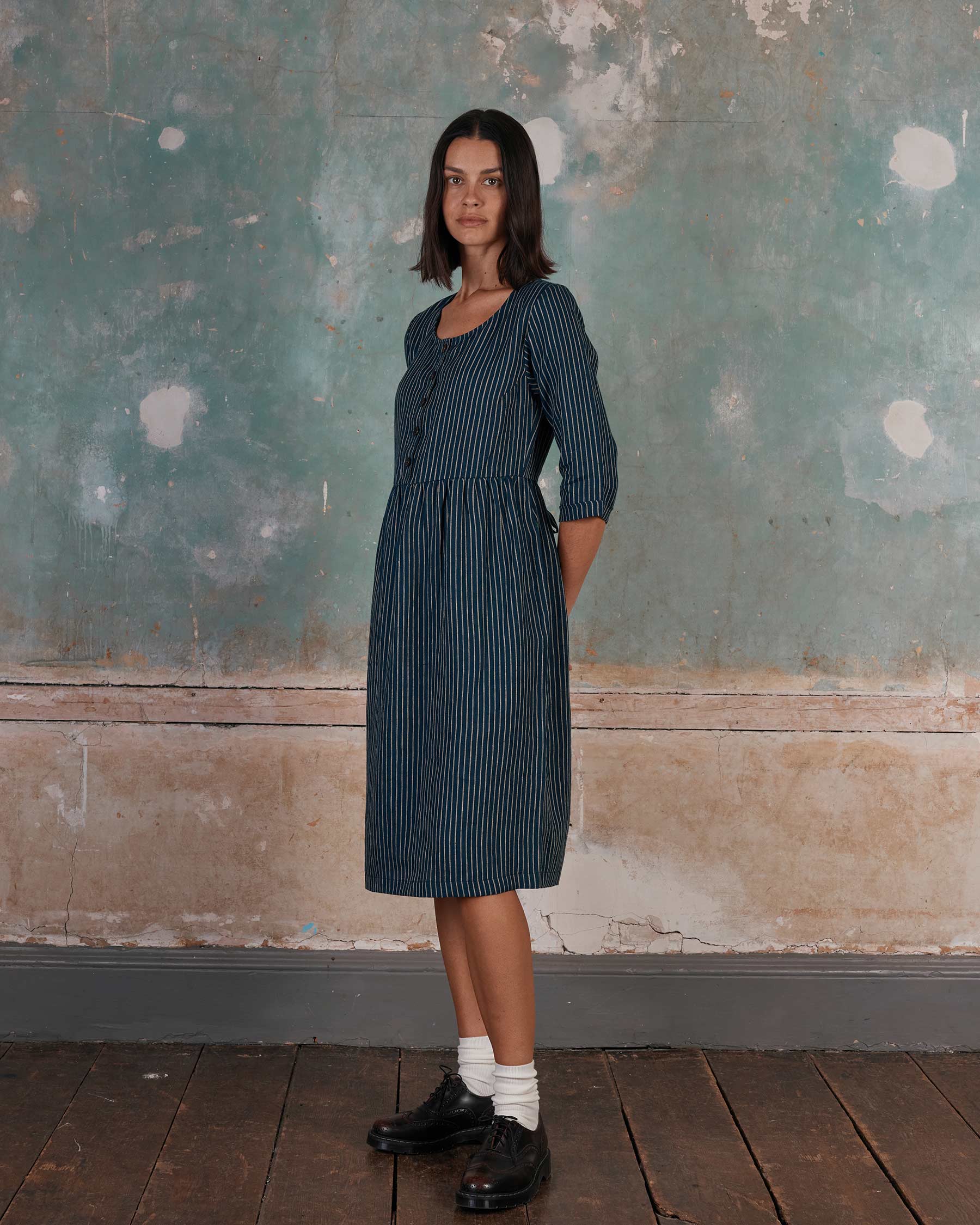 A woman stands indoors against a textured wall, wearing the Pyne and Smith Clothiers No.12 Button Up Linen Midi Dress in Mariner Stripe, made in California. Her hands are behind her back as she looks at the camera.