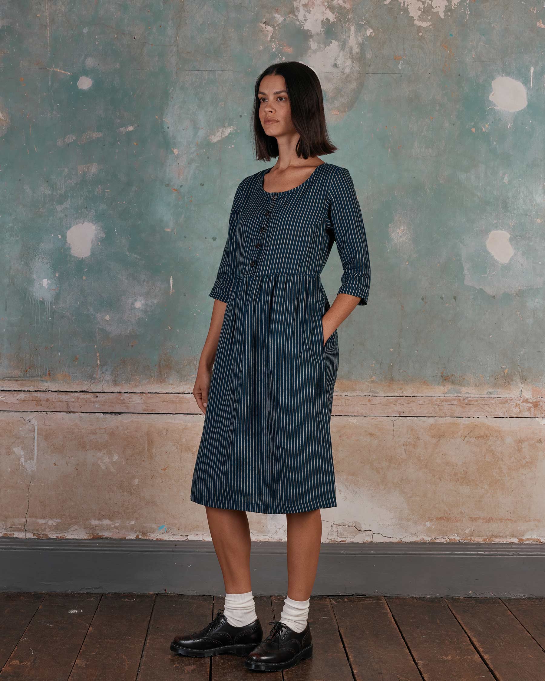A woman with straight dark hair wears the Pyne and Smith Clothiers No.12 Button Up Linen Midi Dress in Mariner Stripe, paired with black shoes and white socks, standing indoors by a weathered wall. Made in California.