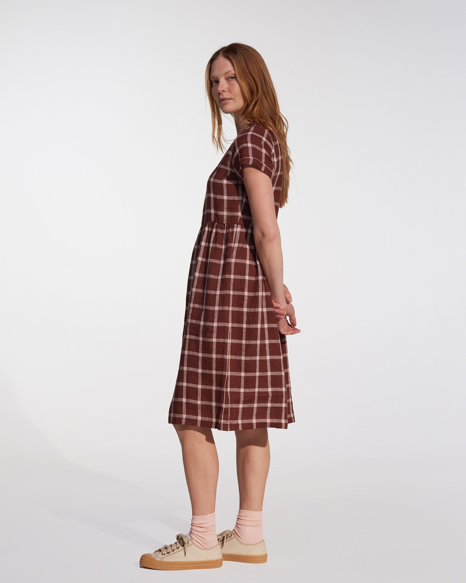 A woman with long red hair stands in profile against a white background, wearing the No.15 Linen Day Dress in Neapolitan by Pyne and Smith Clothiers, paired with light pink socks and beige sneakers.