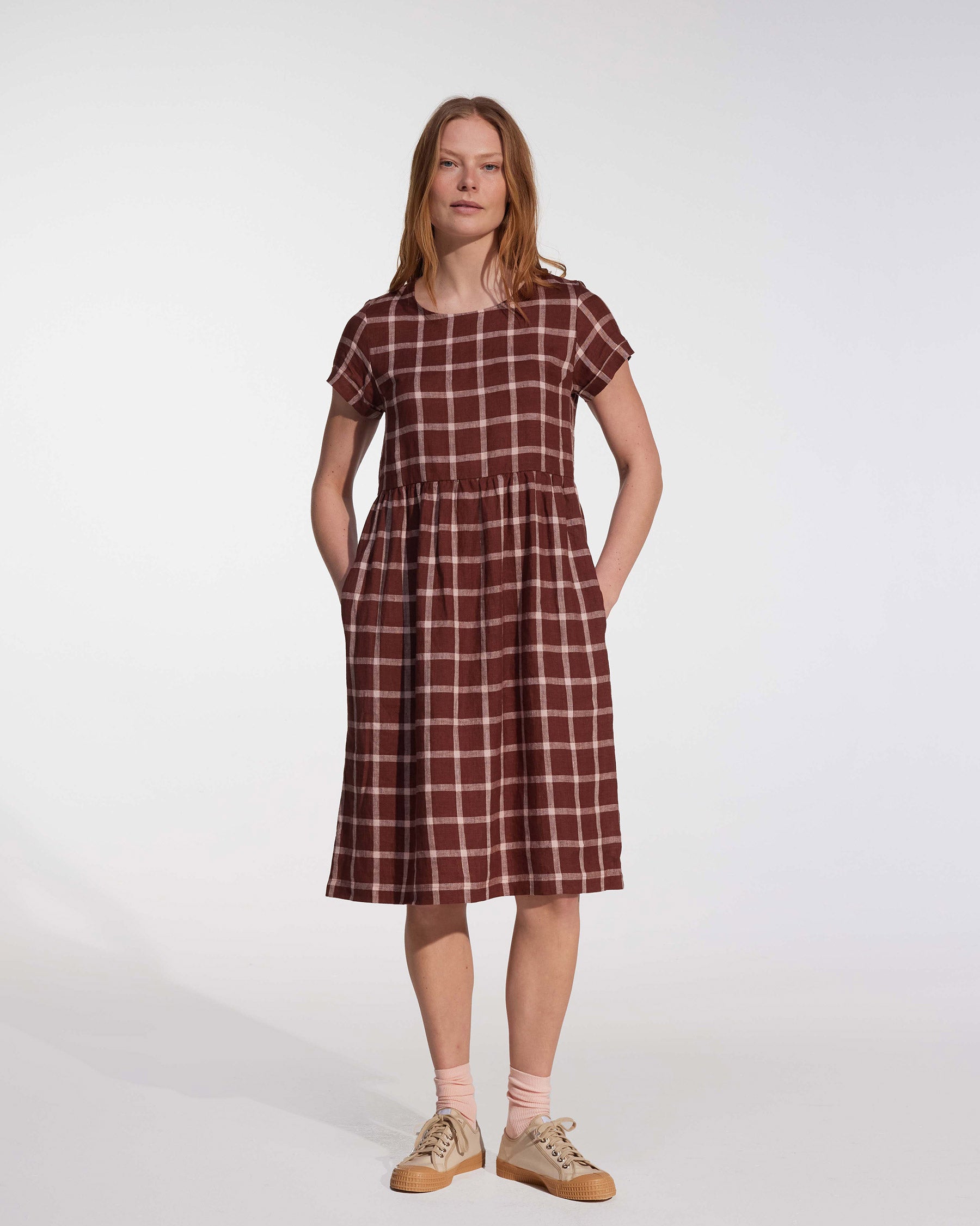 A person stands against a plain background wearing the Pyne and Smith Clothiers No.15 Linen Day Dress in Neapolitan, knee-length with short sleeves, white plaid pattern, and side pockets. Light pink socks and light brown sneakers complete the look, hands in pockets.