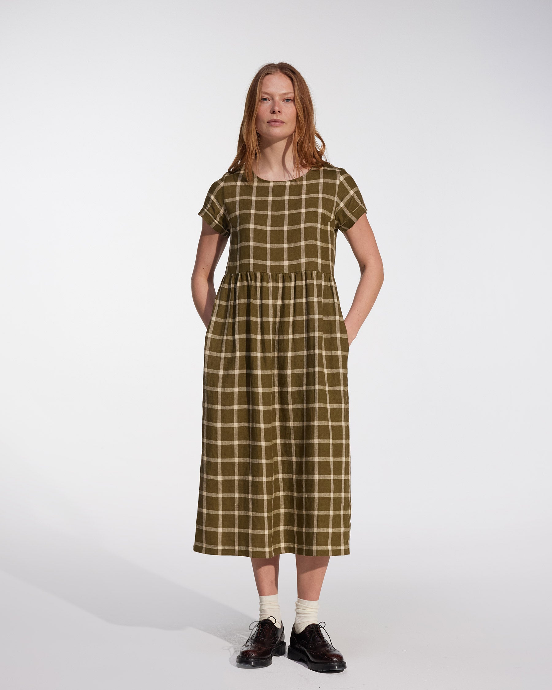 A woman with long hair stands against a plain background wearing the Pyne and Smith Clothiers No.17 Full Length Linen Tea Dress in Celtic Plaid, styled with black shoes and white socks.