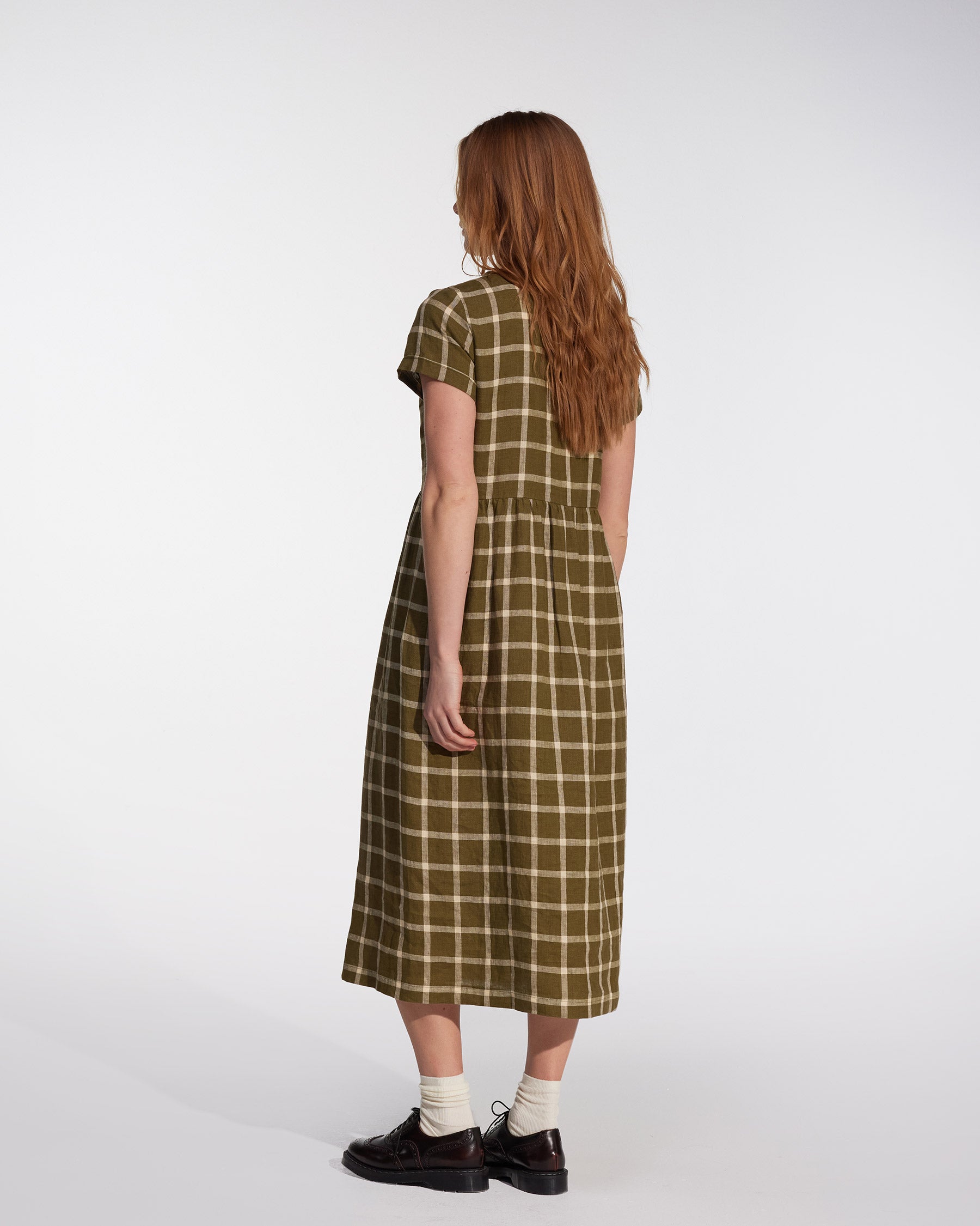 A woman with long, reddish-brown hair stands facing away, wearing the Pyne and Smith Clothiers No.17 Full Length Linen Tea Dress in Celtic Plaid, styled with white socks and black shoes against a plain light background.