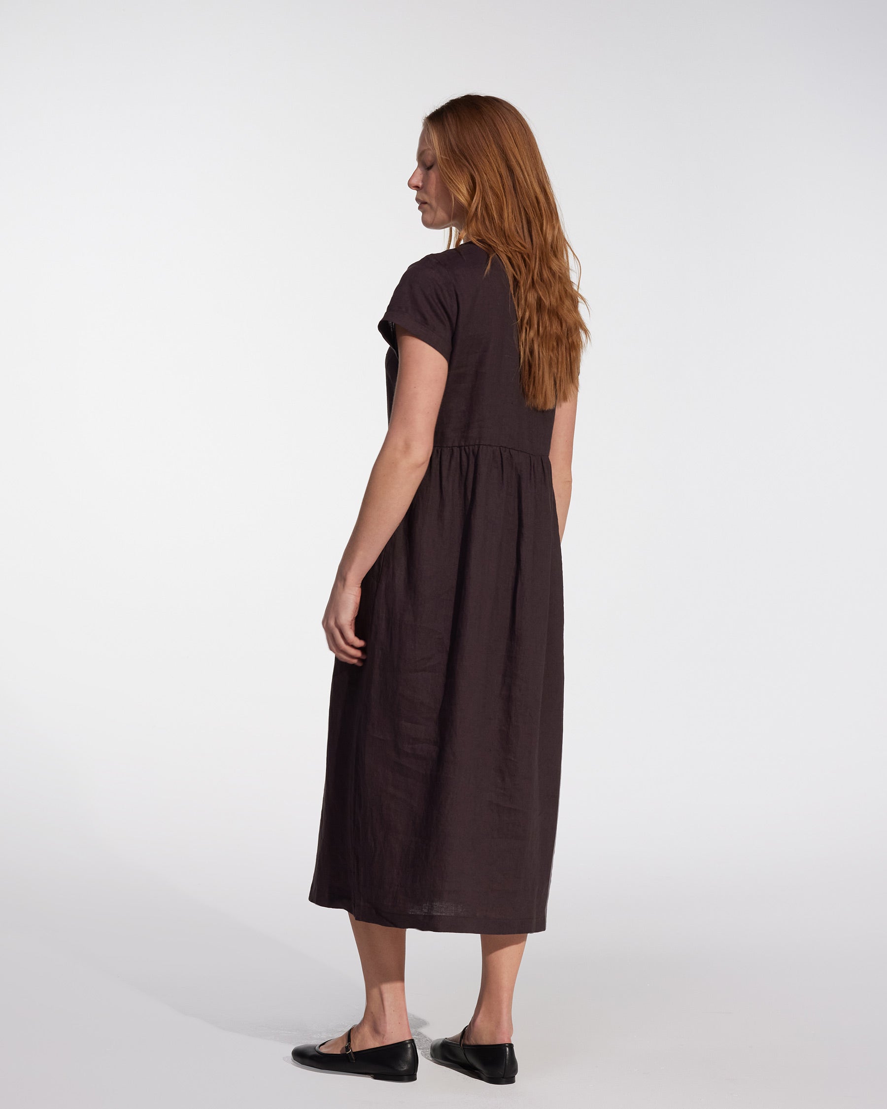 A woman with long red hair wears the Pyne and Smith Clothiers No.17 Full Length Linen Tea Dress in Chocolate Plum, featuring short sleeves and side pockets, styled with black flats, standing sideways against a plain white background.