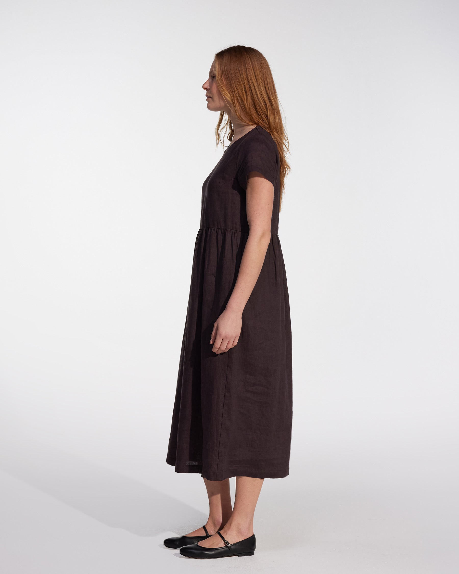 A woman with long, straight hair stands in profile wearing the Pyne and Smith Clothiers No.17 Full Length Linen Tea Dress in Chocolate Plum with side pockets and black flats against a plain white background.