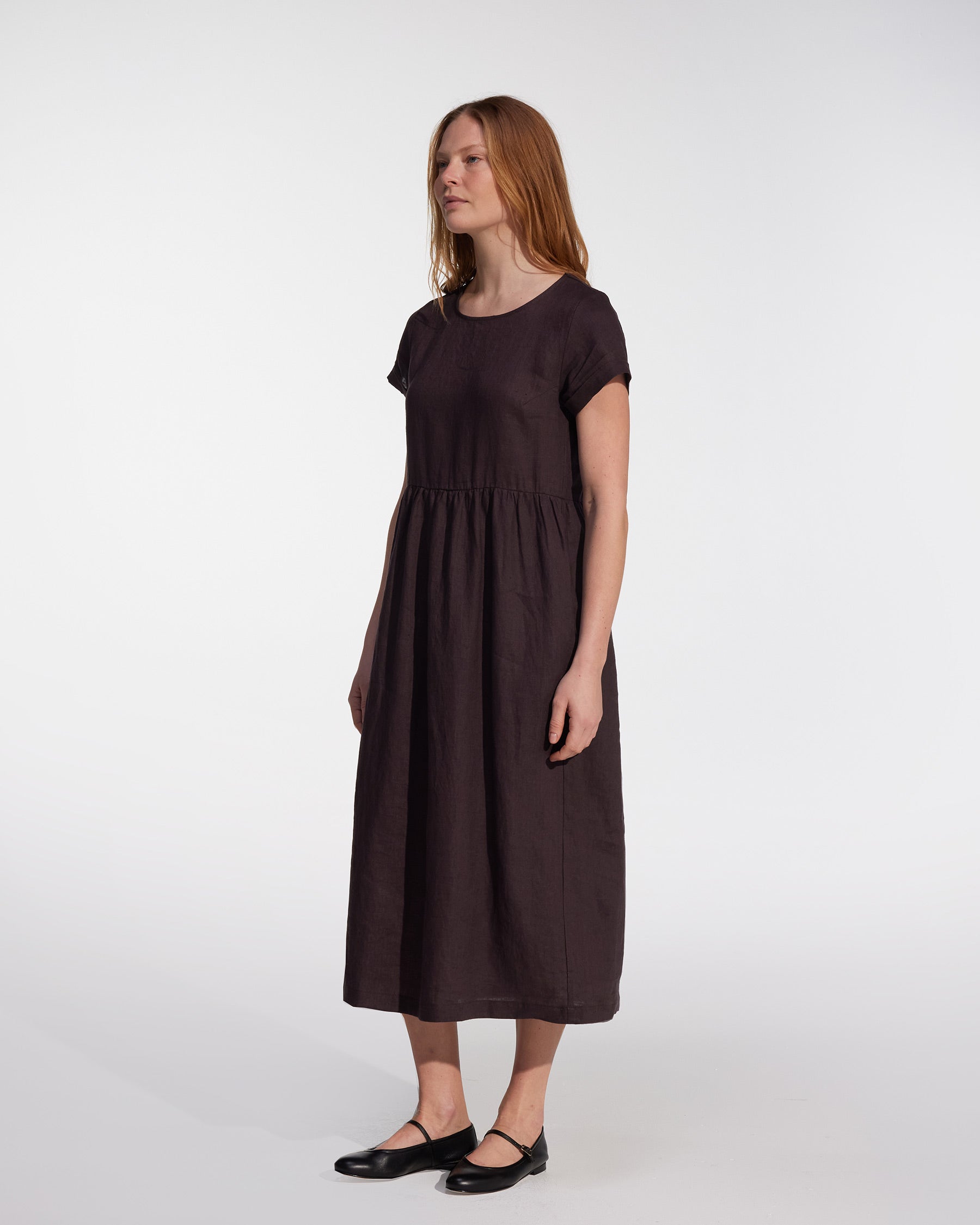 A woman with long, straight hair stands against a white background, wearing Pyne and Smith Clothiers' No.17 Full Length Linen Tea Dress in Chocolate Plum and black shoes.