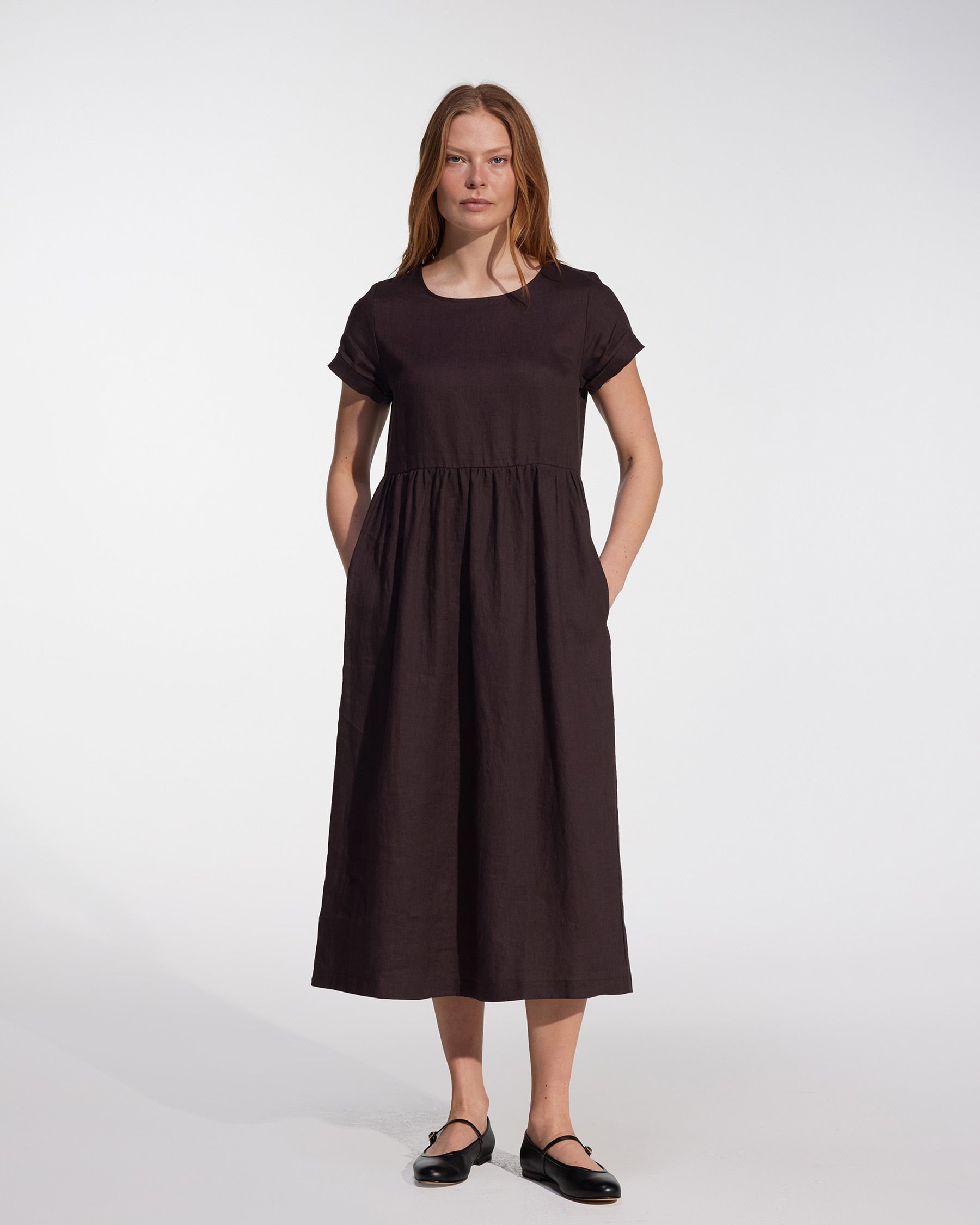 A woman with straight light brown hair wears the Pyne and Smith Clothiers No.17 Full Length Linen Tea Dress in Chocolate Plum, featuring short sleeves, a gathered waist, side pockets, and black flat shoes against a plain background.