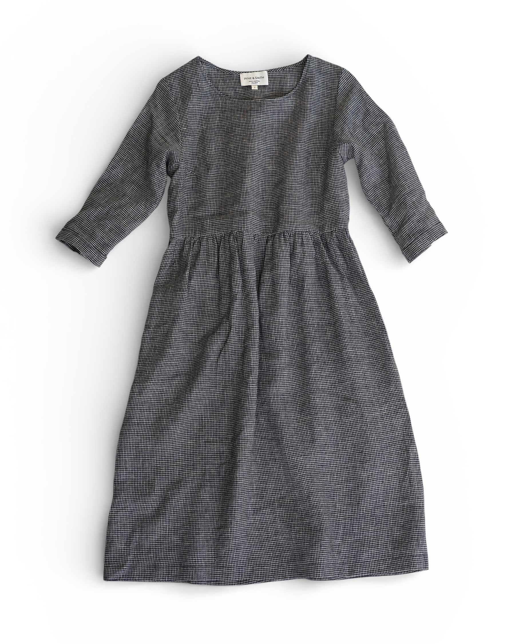 The Pyne and Smith Clothiers No.19 Midi Linen Dress in Brasserie Check features long sleeves, an ankle-length cut, round neckline, fitted bodice, adjustable waist tie, and grey checkered pattern, shown laid flat on a white background.