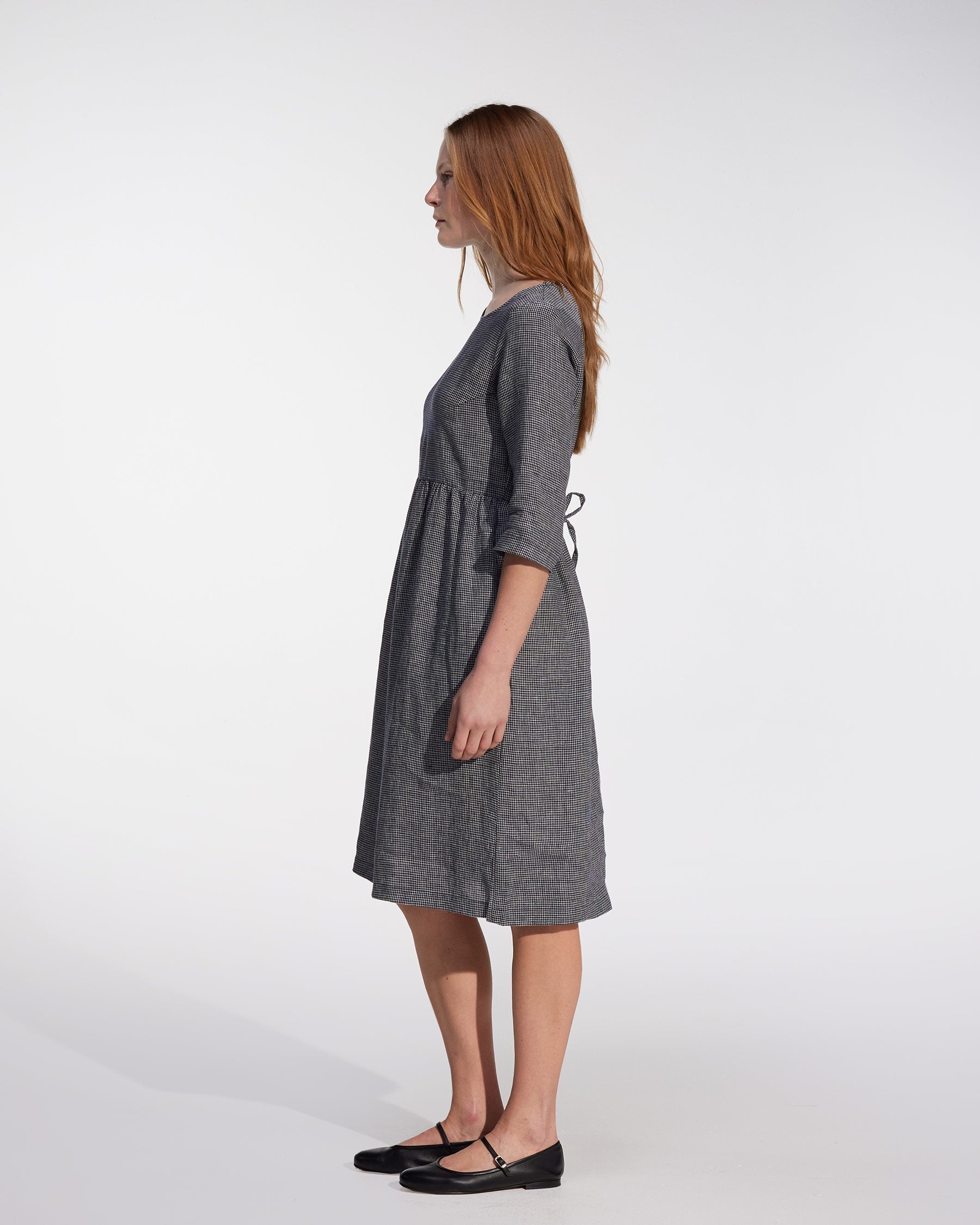 A woman with long, straight hair stands in profile against a white background, wearing the Pyne and Smith Clothiers No.19 Midi Linen Dress in Brasserie Check and black flat shoes.