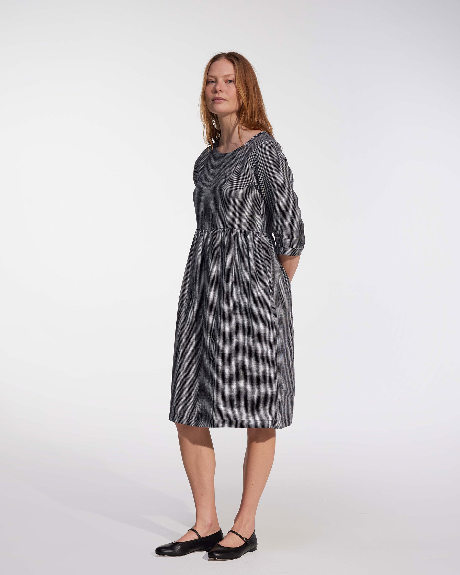 A woman with long, light brown hair stands against a white background wearing the Pyne and Smith Clothiers No.19 Midi Linen Dress in Brasserie Check, featuring an adjustable waist tie and knee-length fit for a classic, elegant look. Made in California.