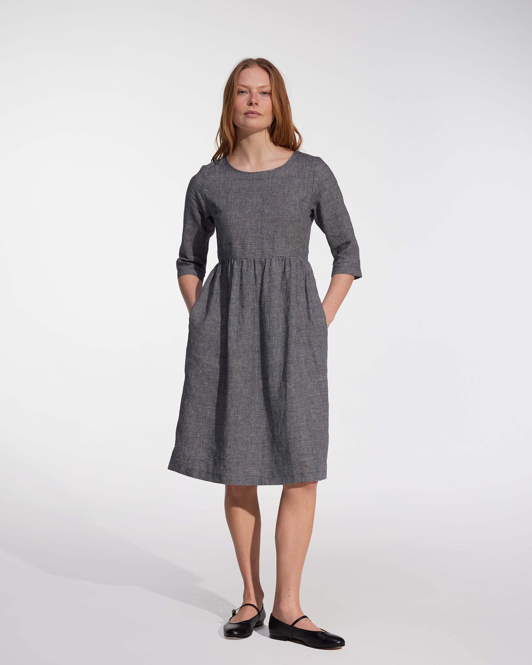 A woman with light brown hair wears the Pyne and Smith Clothiers No.19 Midi Linen Dress in Brasserie Check, featuring three-quarter sleeves, adjustable waist tie, and pockets, styled with black flats. Made in California against a white background.