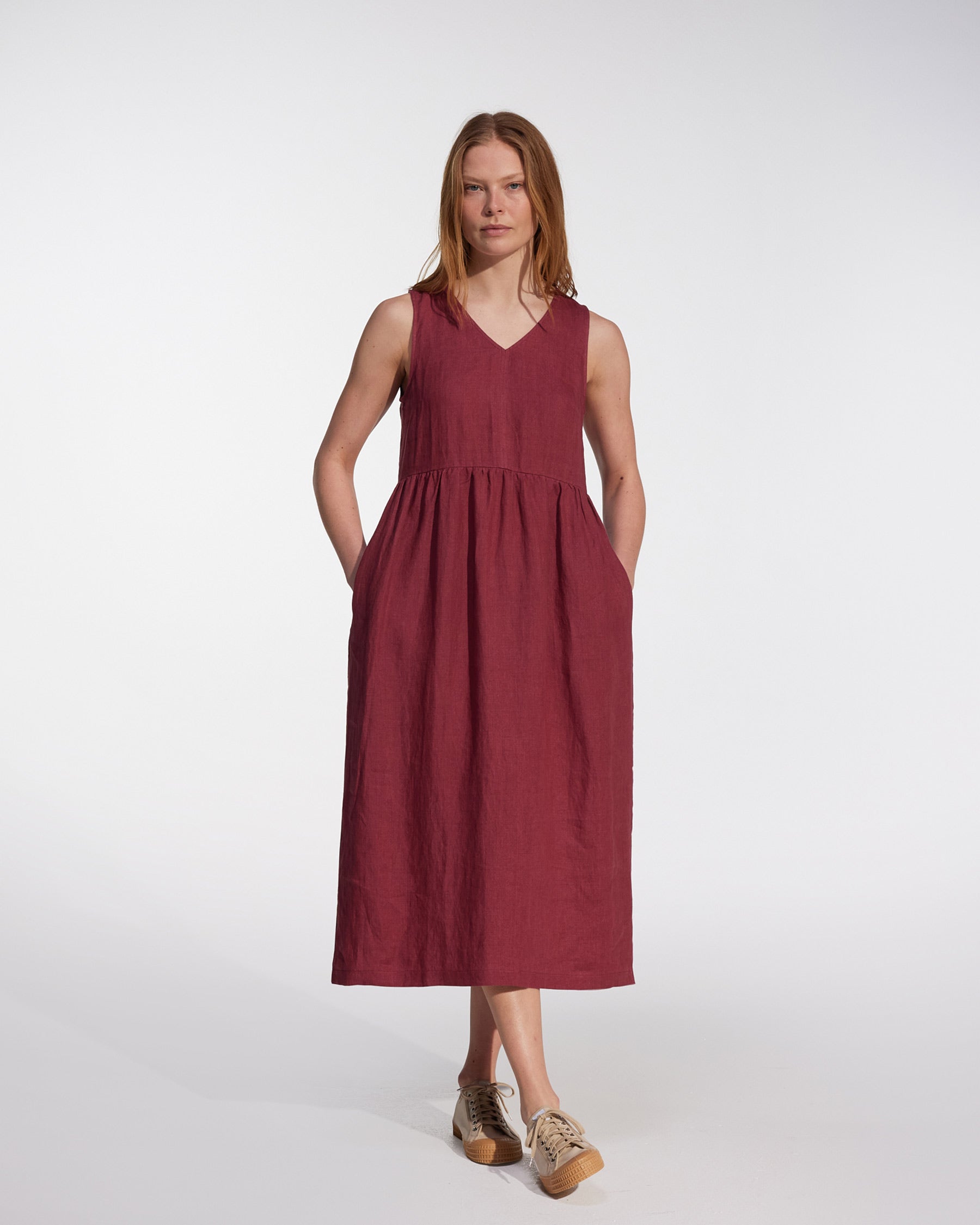 A woman with straight light brown hair and a neutral expression wears the No.21 V Neck Sleeveless Linen Dress in Roan by Pyne and Smith Clothiers, styled with light sneakers, standing against a plain white background.