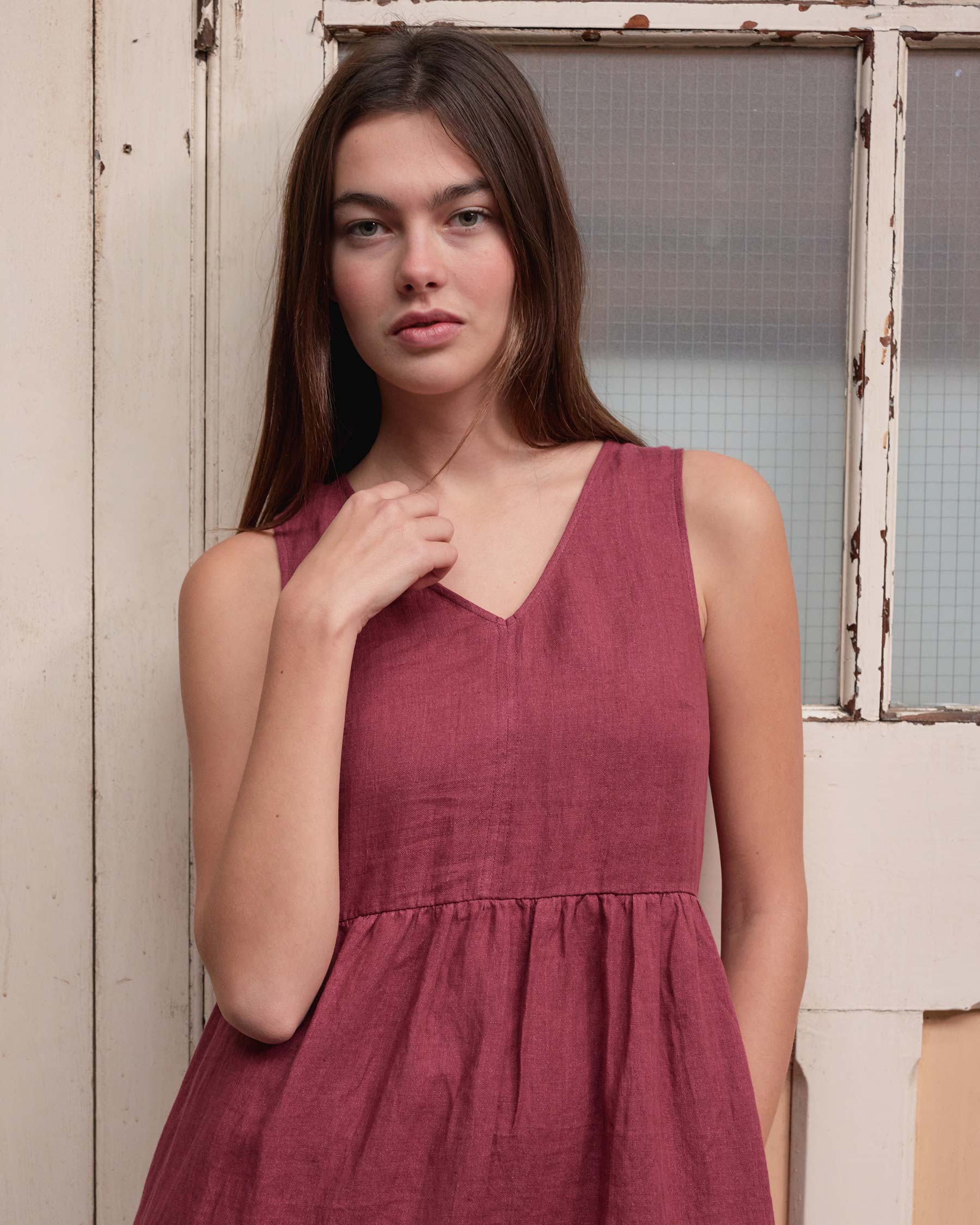 A woman with long brown hair wears the Pyne and Smith Clothiers No.21 V Neck Sleeveless Linen Dress in Roan, posing by a rustic window and door, looking at the camera with a neutral expression.