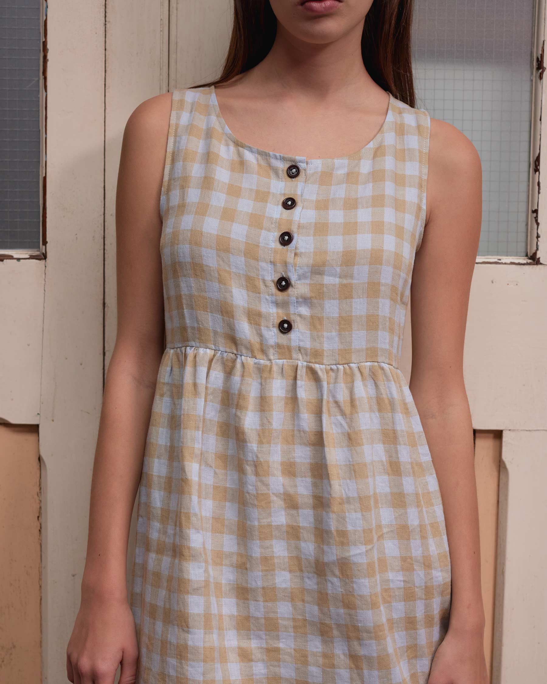 A woman, her face partly out of frame, stands before a wooden door in the No.24 Sleeveless, Button Up Linen Dress in St. Agnes Check by Pyne and Smith Clothiers, made from European flax linen. Only her upper body is visible.