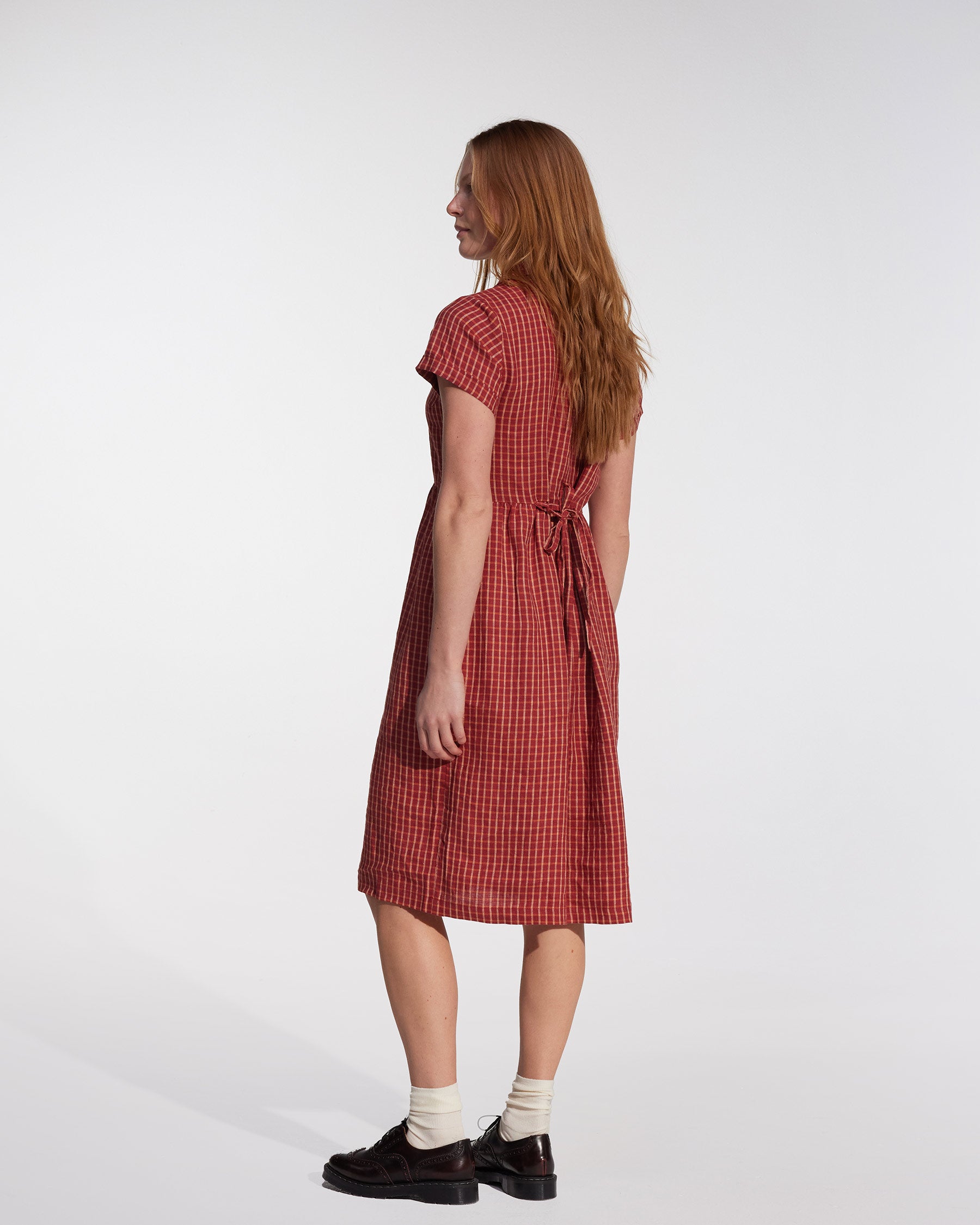 A woman with long reddish hair stands sideways, wearing the No.30 Collared Linen Dress in Pomodoro Plaid by Pyne and Smith Clothiers, paired with white socks and dark shoes against a plain light background.