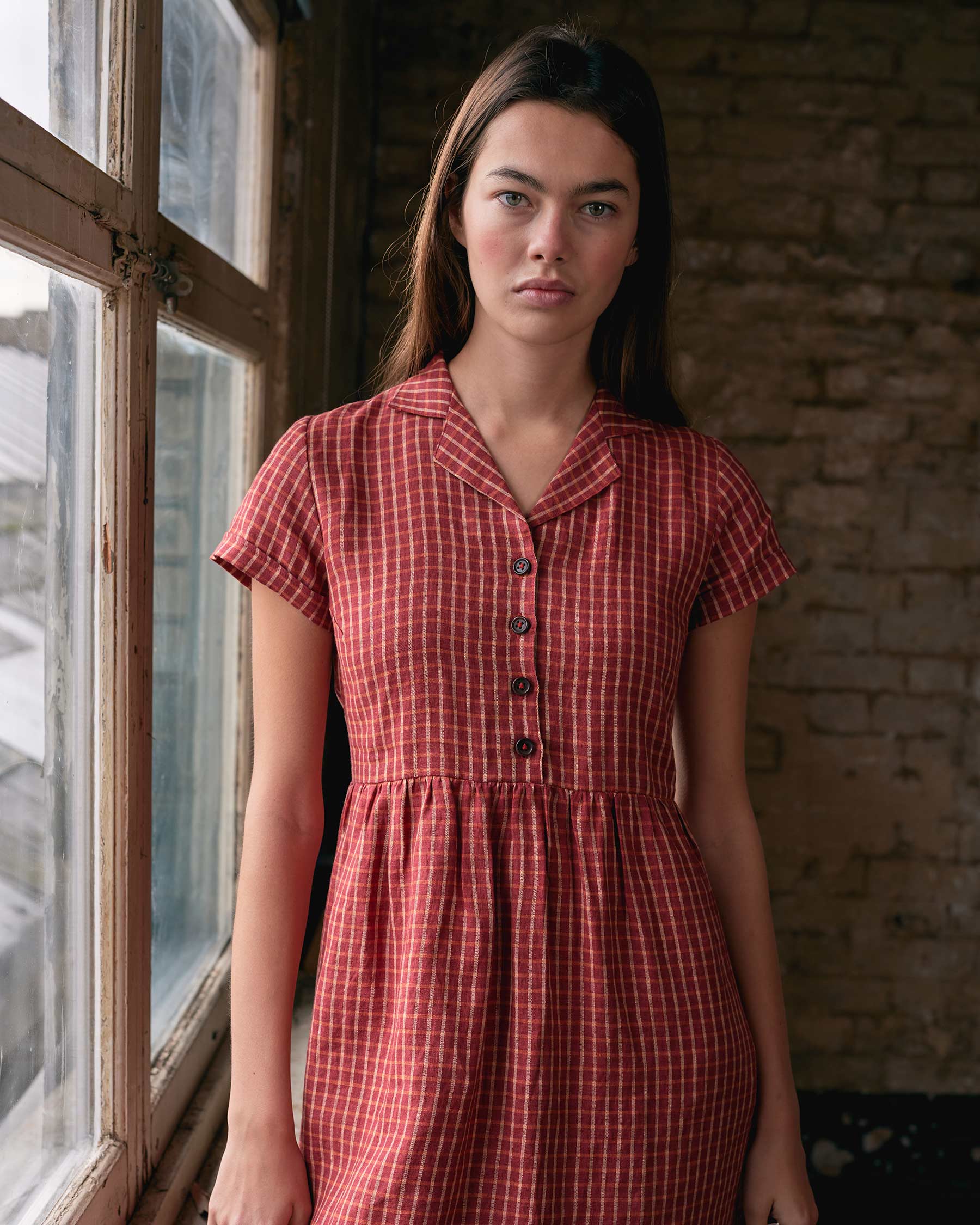 A woman with long brown hair wears the No.30 Collared Linen Dress in Pomodoro Plaid by Pyne and Smith Clothiers, standing indoors by a window and facing the camera against an exposed brick wall.