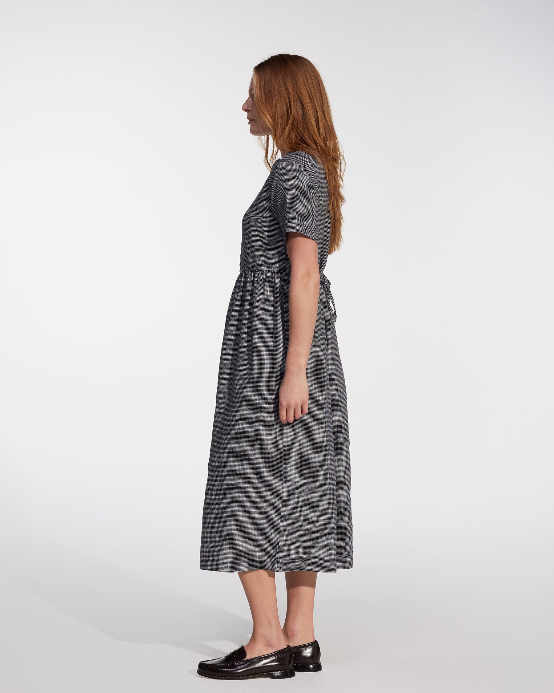 A woman with long, light brown hair stands in profile wearing the Pyne and Smith Clothiers No.31 V Neck, Full Length Linen Dress in Brasserie Check with black loafers against a plain, light background. Made in California.