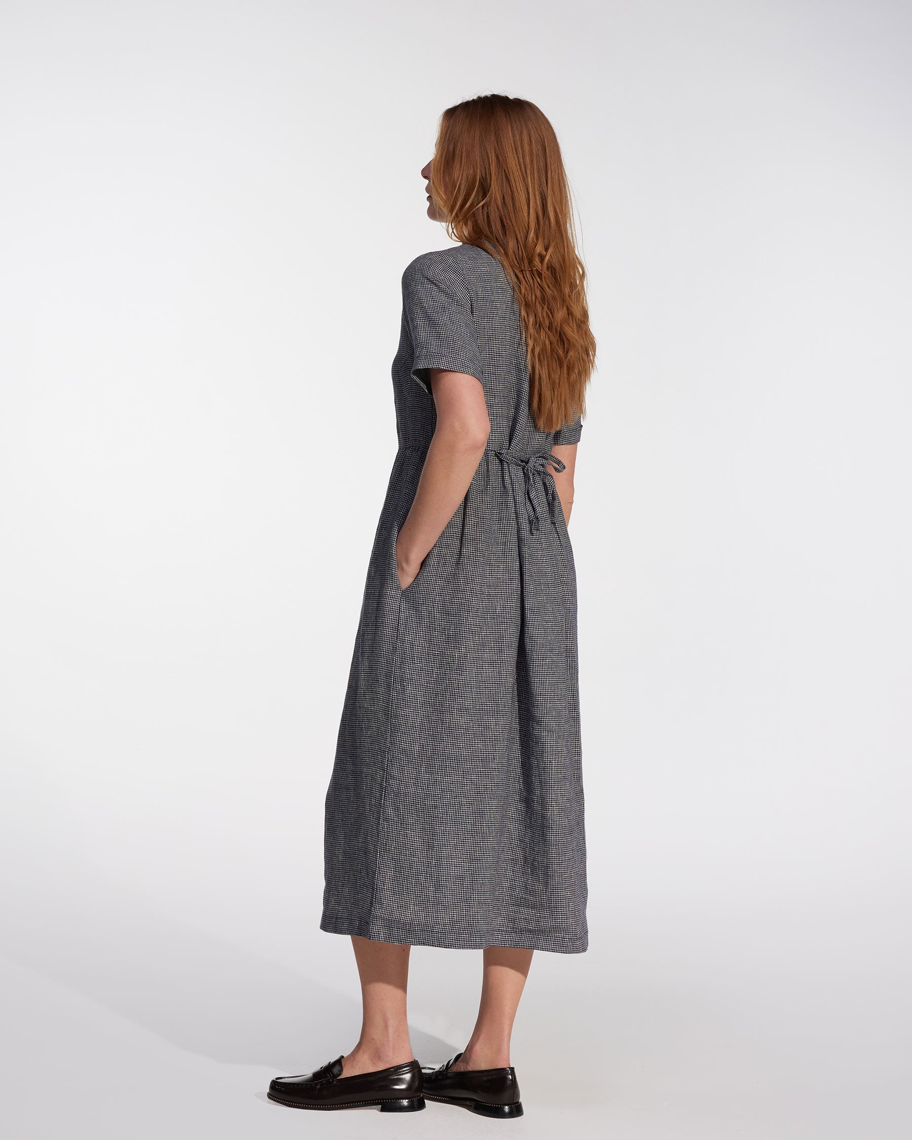 A woman with long auburn hair stands sideways in the Pyne and Smith Clothiers No.31 V Neck, Full Length Linen Dress in Brasserie Check, featuring side pockets and a tied back, paired with black loafers against a plain white background.