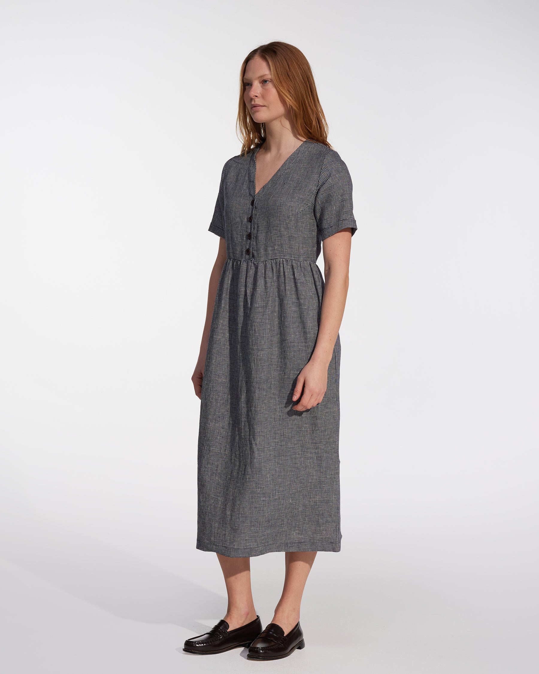 A woman with long, straight hair wears the No.31 V Neck, Full Length Linen Dress in Brasserie Check by Pyne and Smith Clothiers with black shoes, standing slightly to the side against a plain white background.