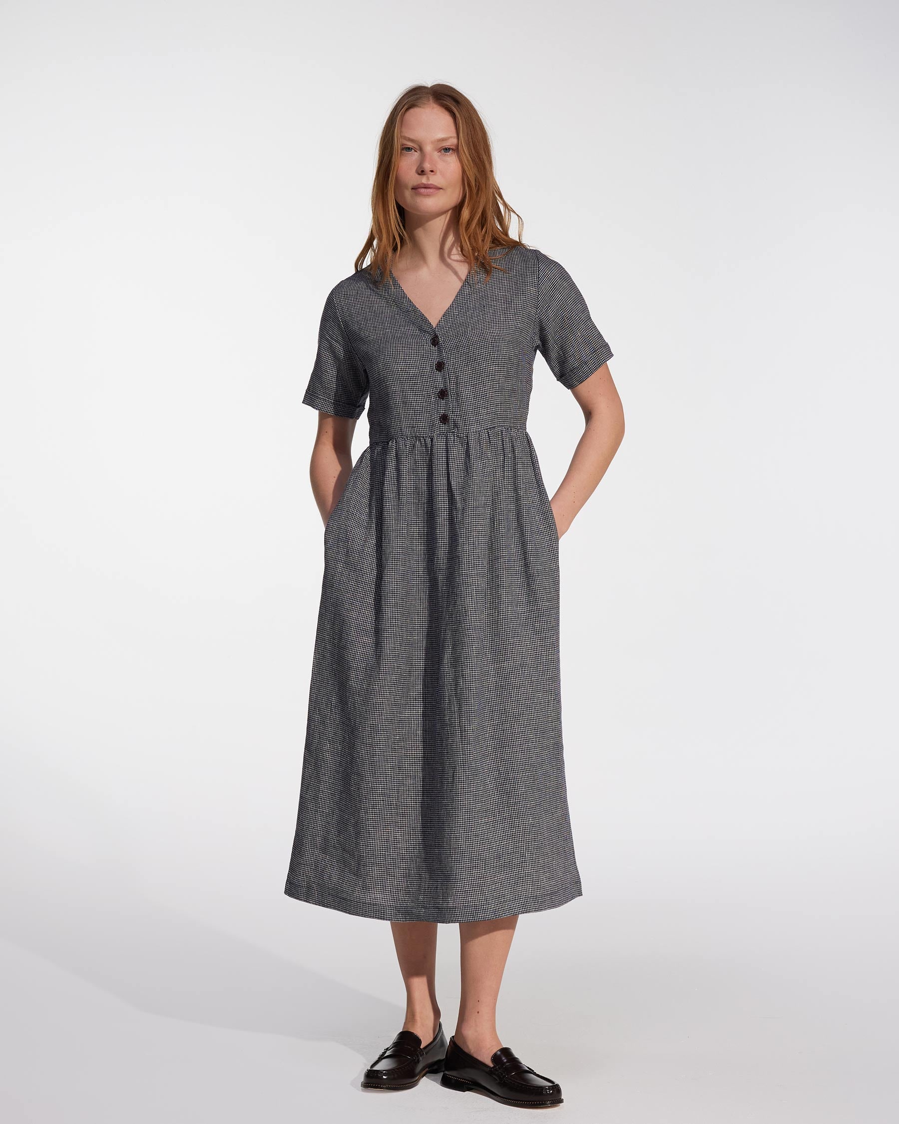 A woman with straight, light brown hair and a neutral expression wears the Pyne and Smith Clothiers No.31 V Neck, Full Length Linen Dress in Brasserie Check, standing against a white background with black shoes.