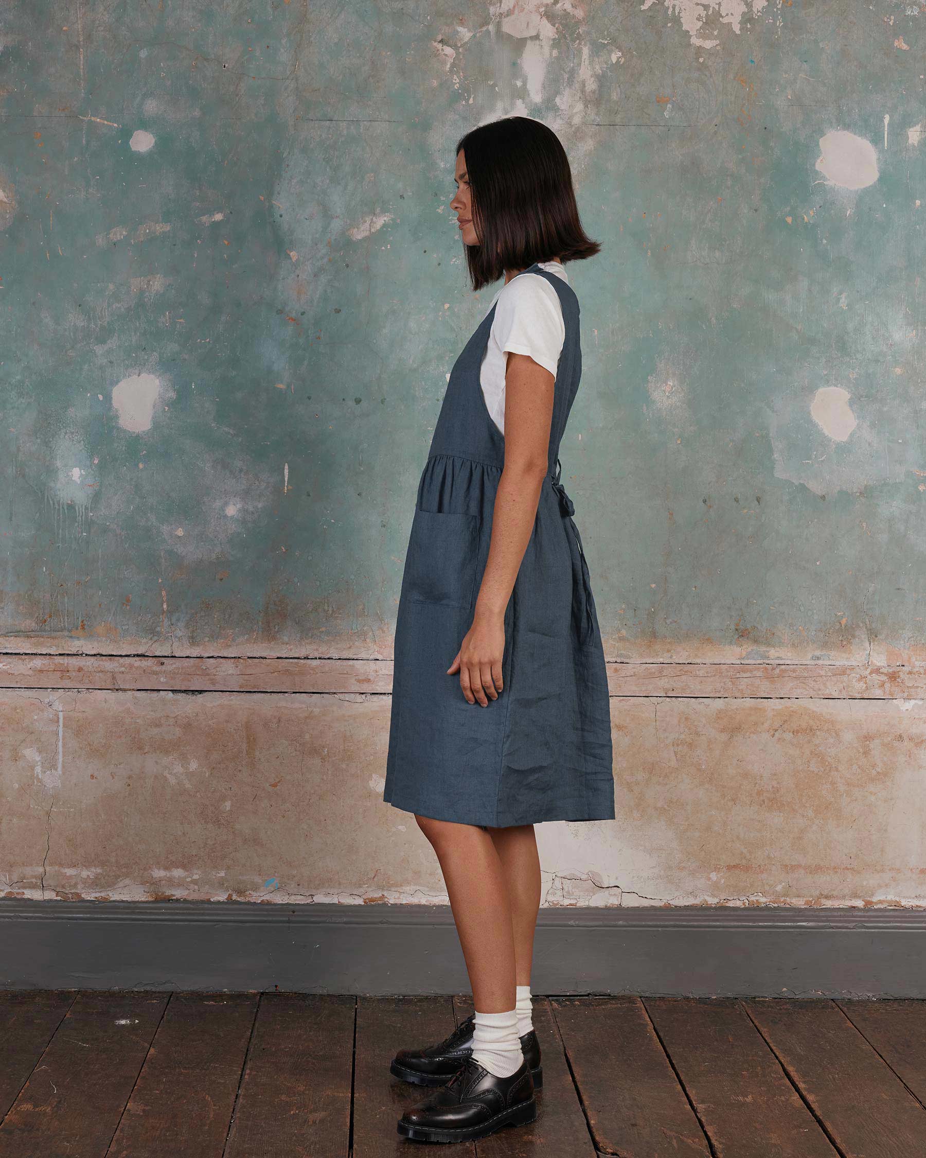 A person with chin-length dark hair stands in profile by a textured wall, wearing a white tee and Pyne and Smith Clothiers’ No.32 The Pinafore Dress in Vintage Indigo, paired with white socks and black shoes on a dark wooden floor.