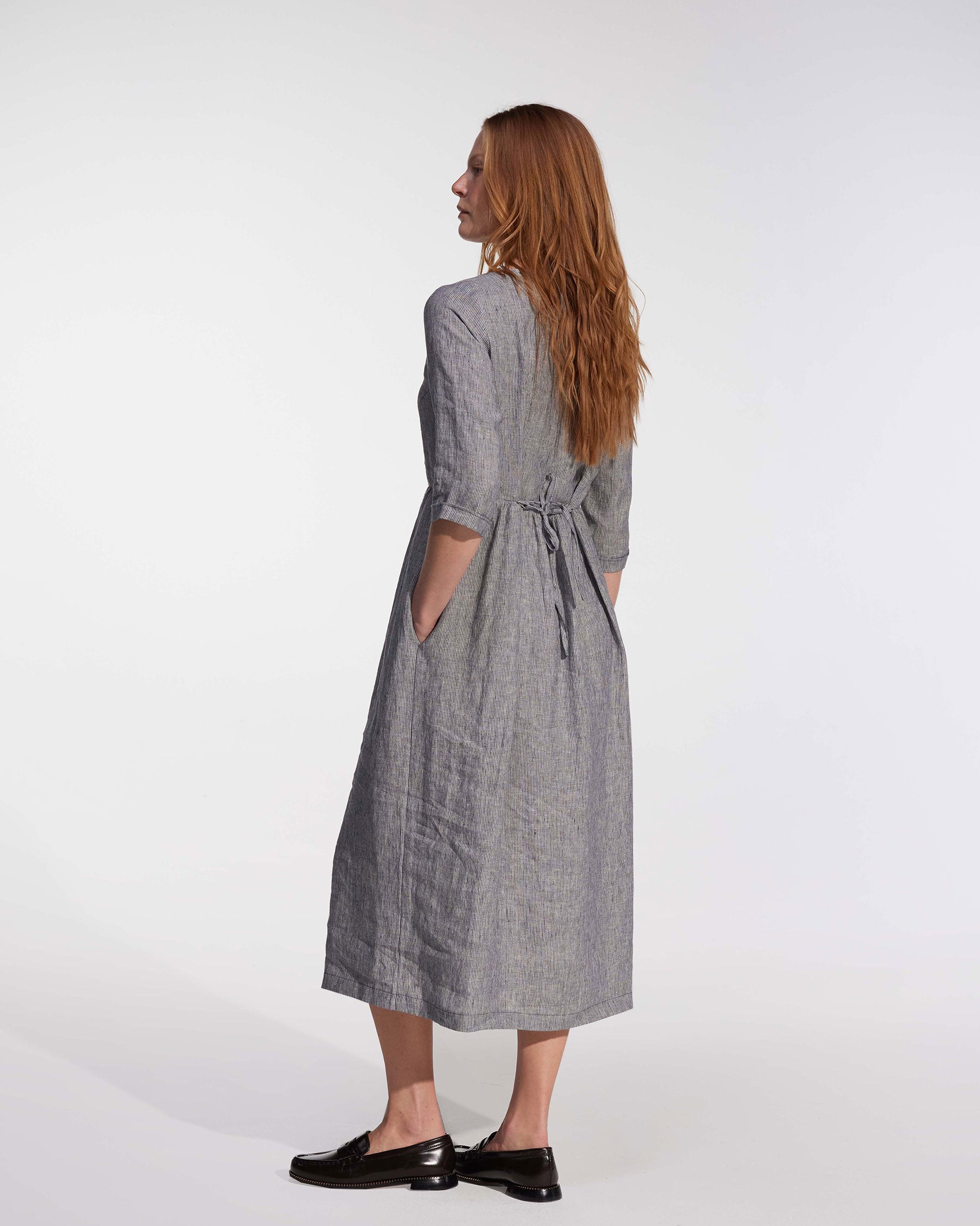 A woman with long, reddish-brown hair stands in profile wearing the No.41 Full Length Linen Day Dress in Cooke's Stripe by Pyne and Smith Clothiers, featuring pockets and an adjustable waist tie, paired with black loafers against a light background. Made in California.