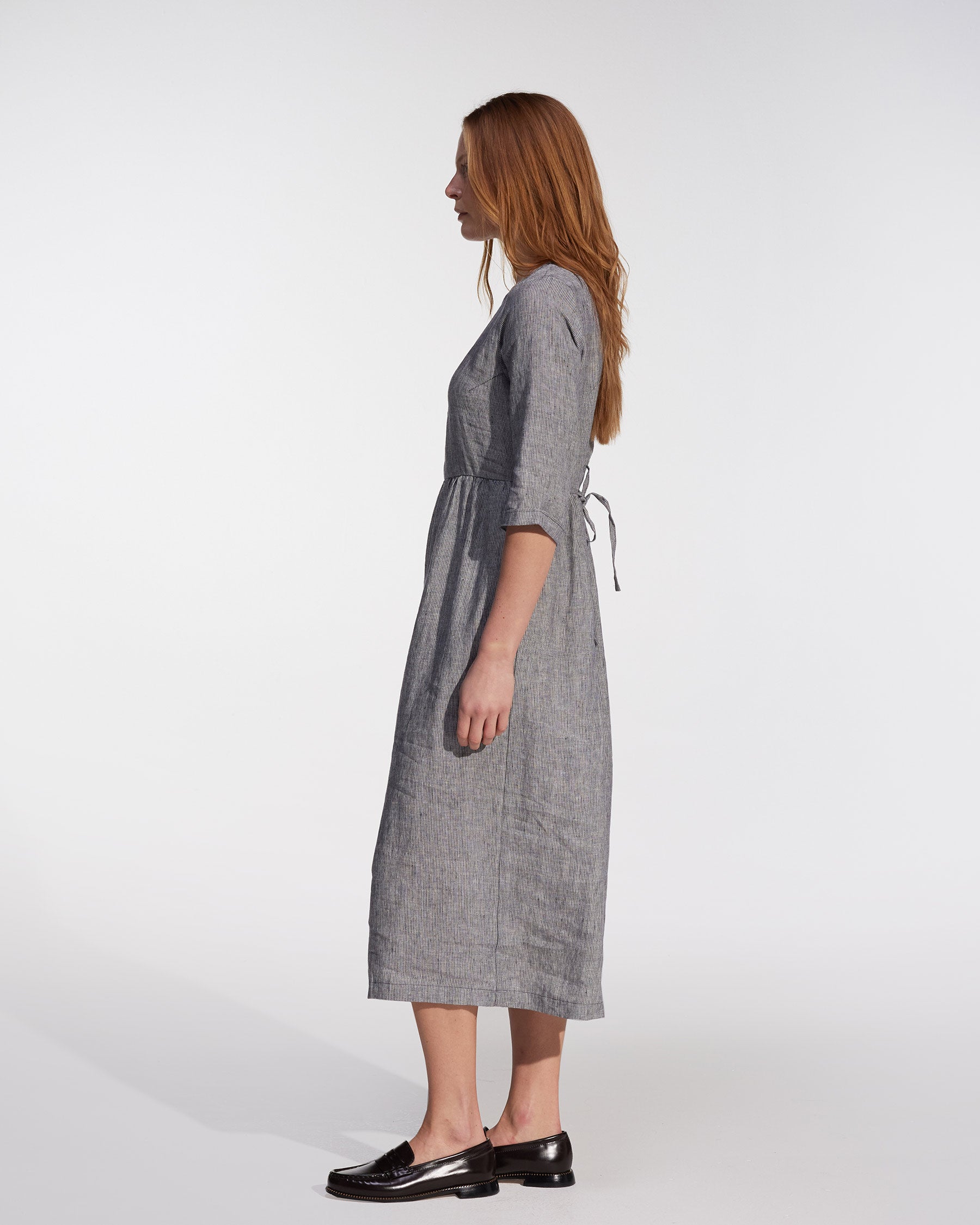 A person with long reddish-brown hair stands in profile wearing the Pyne and Smith Clothiers No.41 Full Length Linen Day Dress in Cooke's Stripe and black loafers, set against a plain, light background.