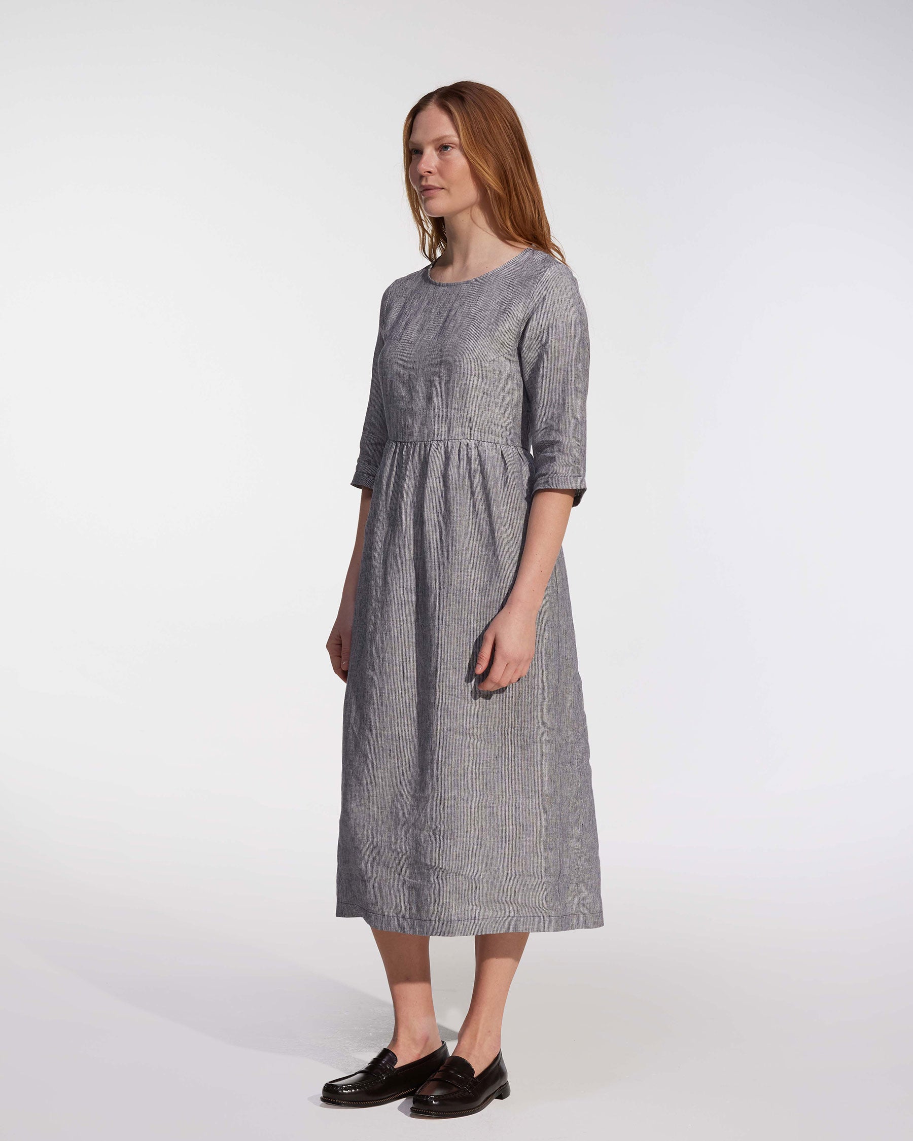 A person with long, straight, reddish hair wears the No.41 Full Length Linen Day Dress in Cooke's Stripe by Pyne and Smith Clothiers, paired with black loafers, standing against a plain white background.