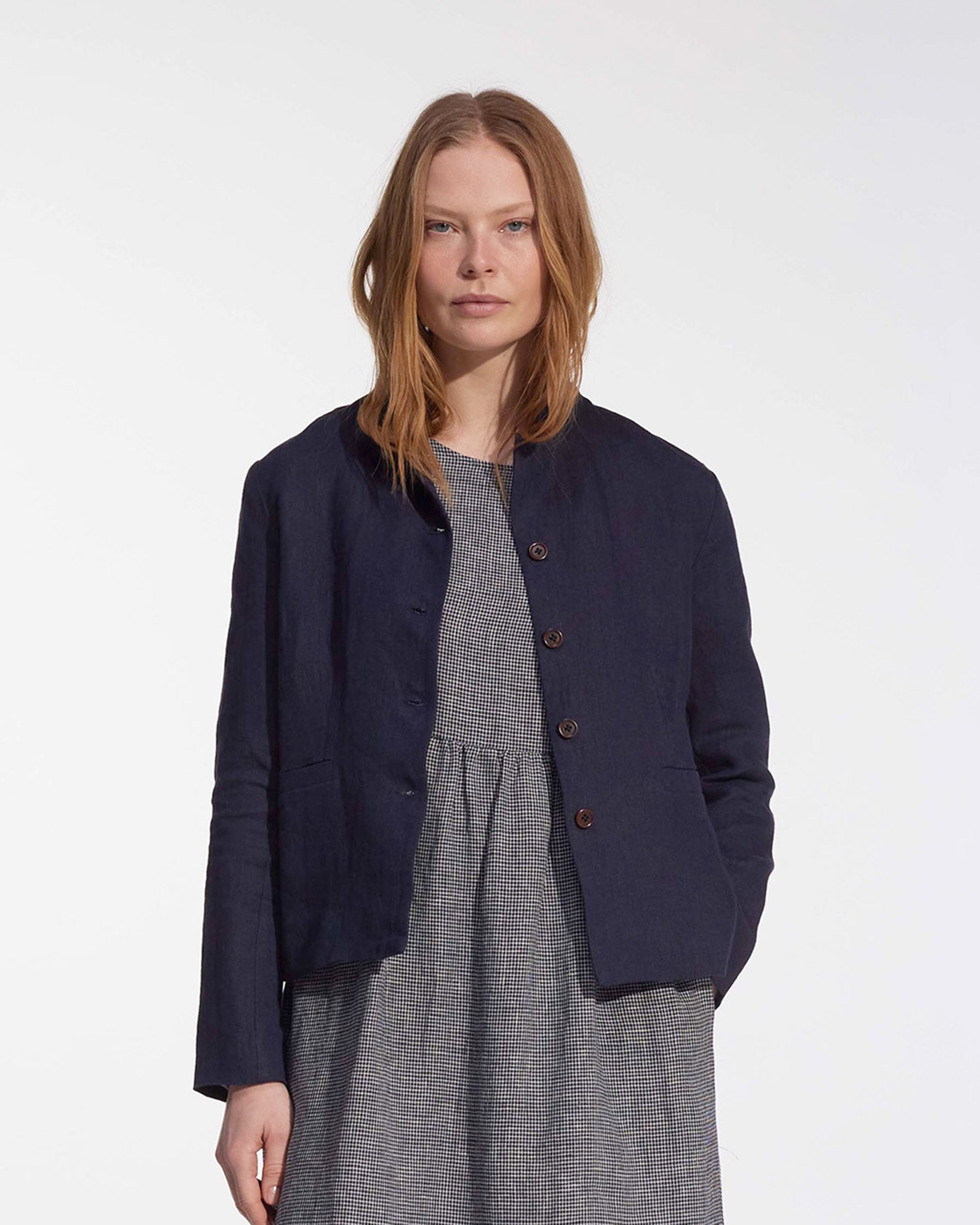 A woman with straight, light brown hair wears the Pyne and Smith Clothiers Tailored Linen Jacket No.10 in Mariner Blue over a gray and white checkered dress, standing against a plain white background.