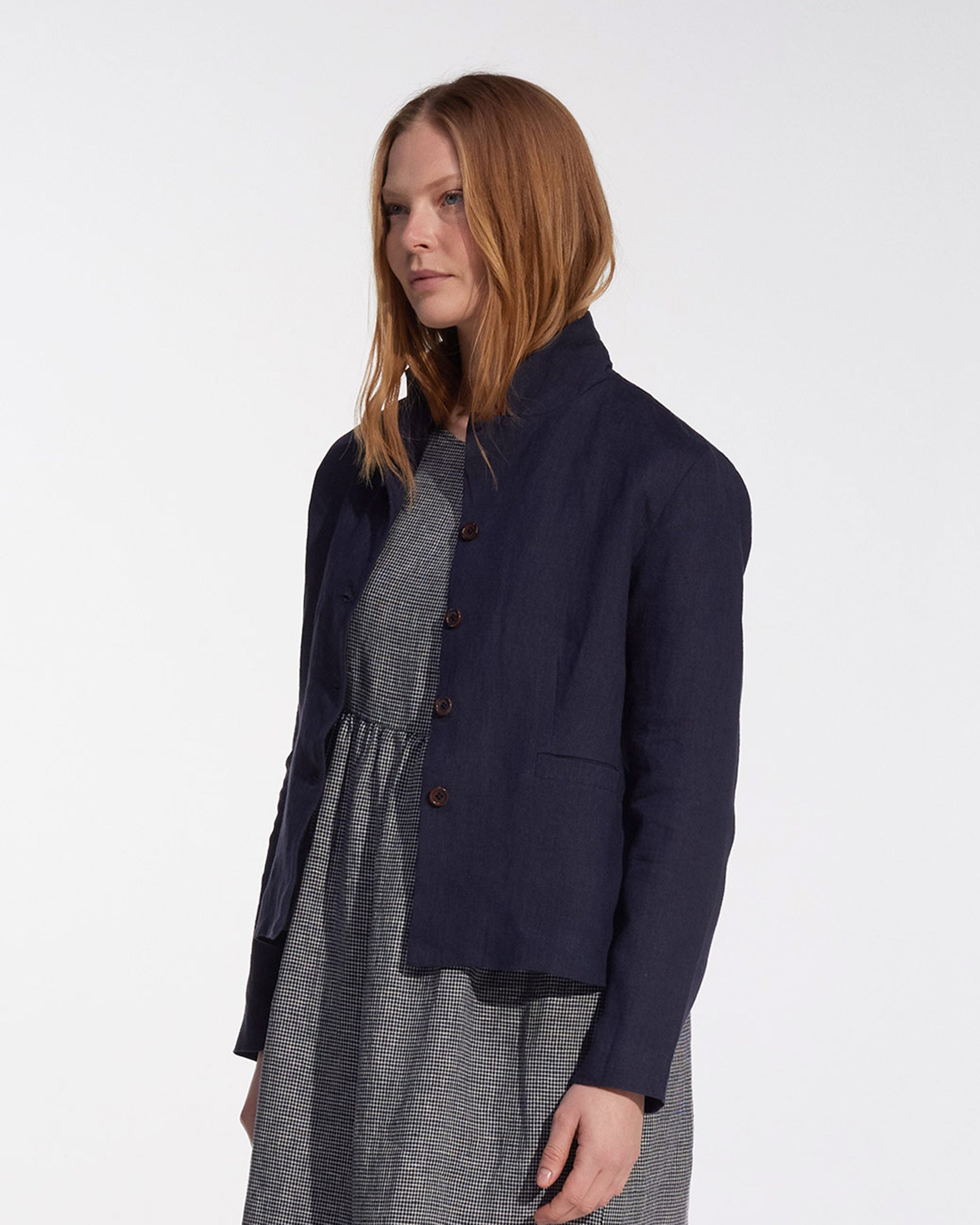 A woman with straight, shoulder-length light brown hair wears the Pyne and Smith Clothiers Tailored Linen Jacket No.10 in Mariner Blue over a gray patterned dress, standing in front of a plain white background.