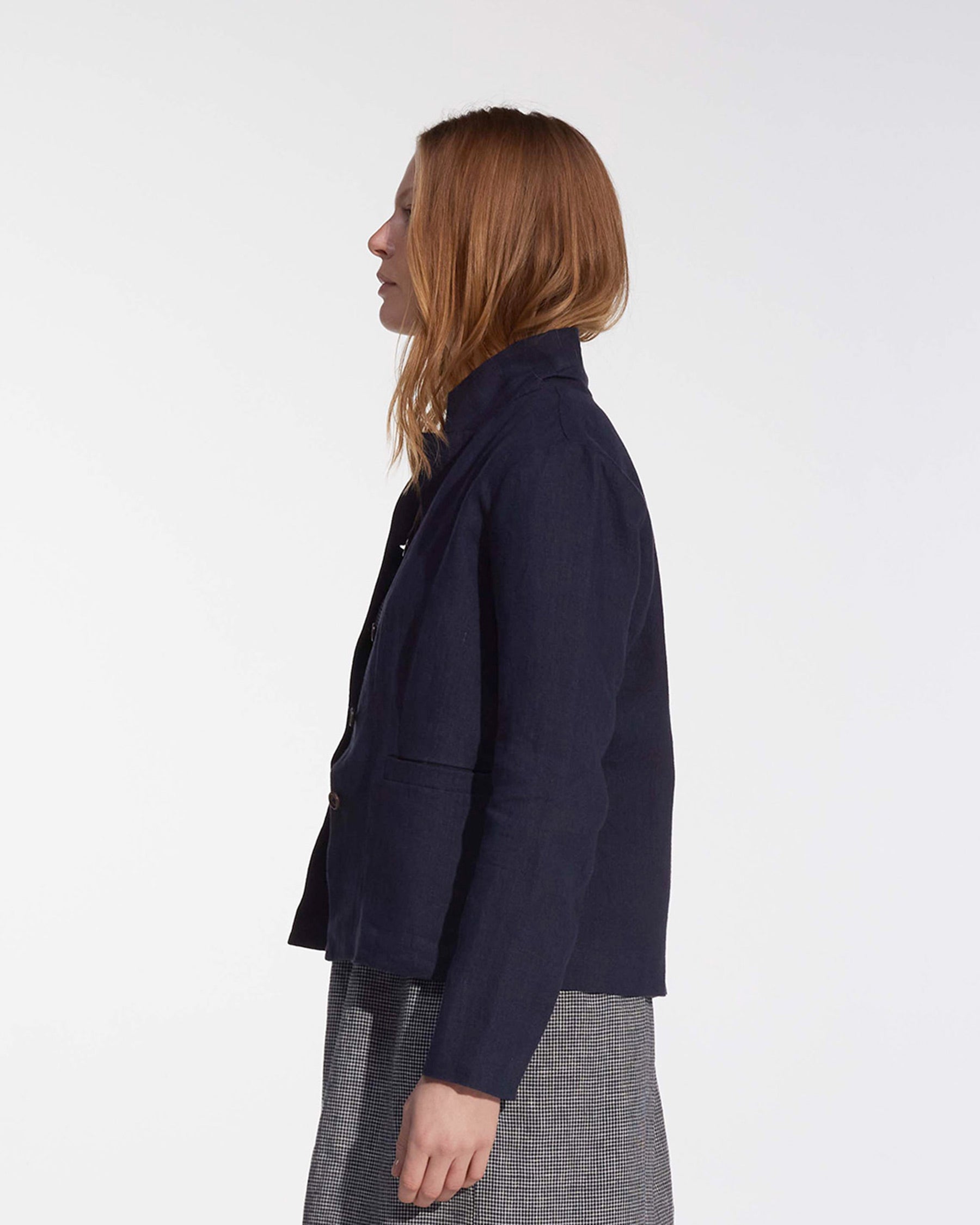A woman with straight, light brown hair stands in profile against a white background, wearing the Pyne and Smith Clothiers Tailored Linen Jacket No.10 in Mariner Blue and a gray plaid skirt.