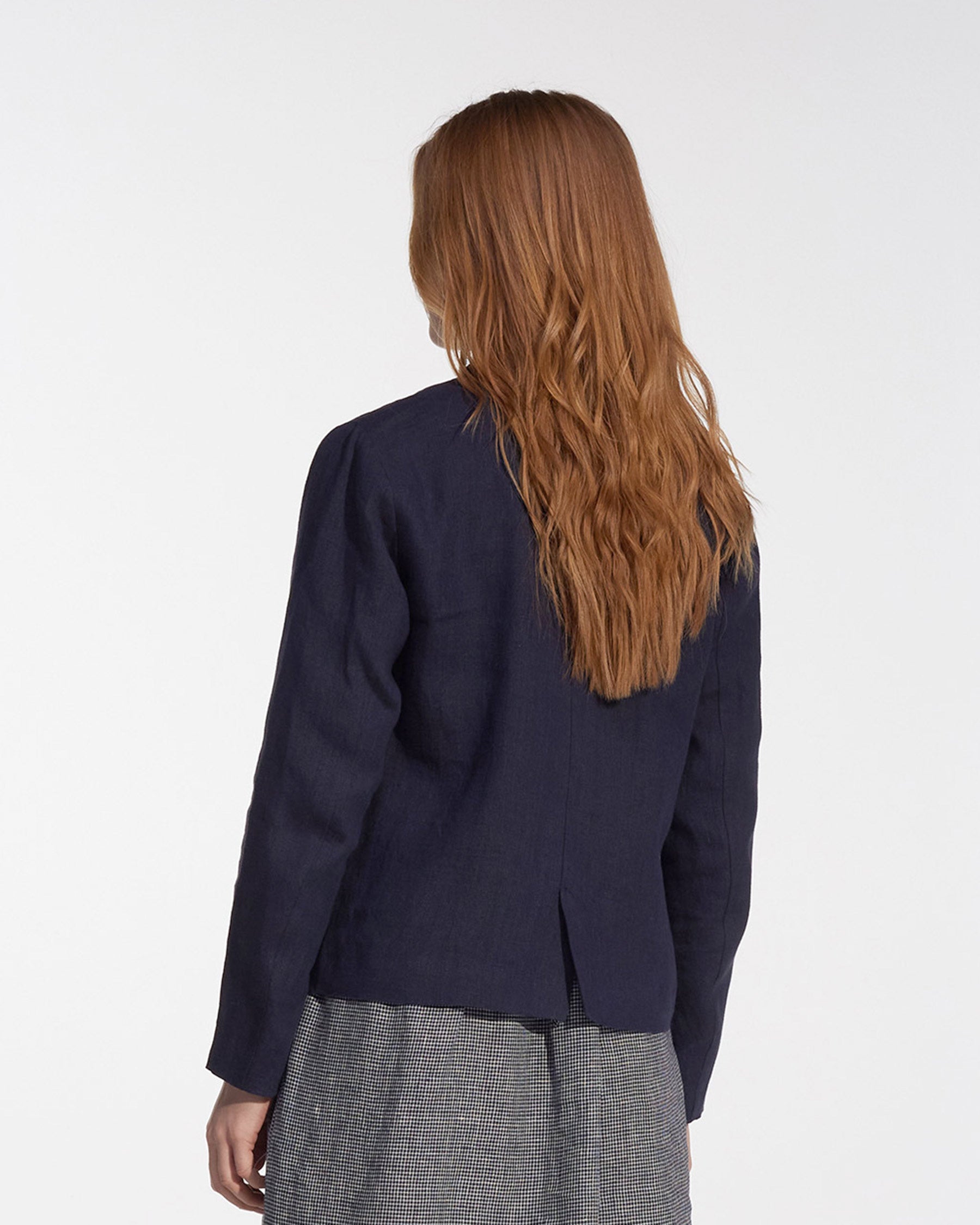 A person with long, wavy, light brown hair is shown from behind in a Pyne and Smith Clothiers Tailored Linen Jacket No.10 in Mariner Blue and a gray checkered skirt against a plain white background.