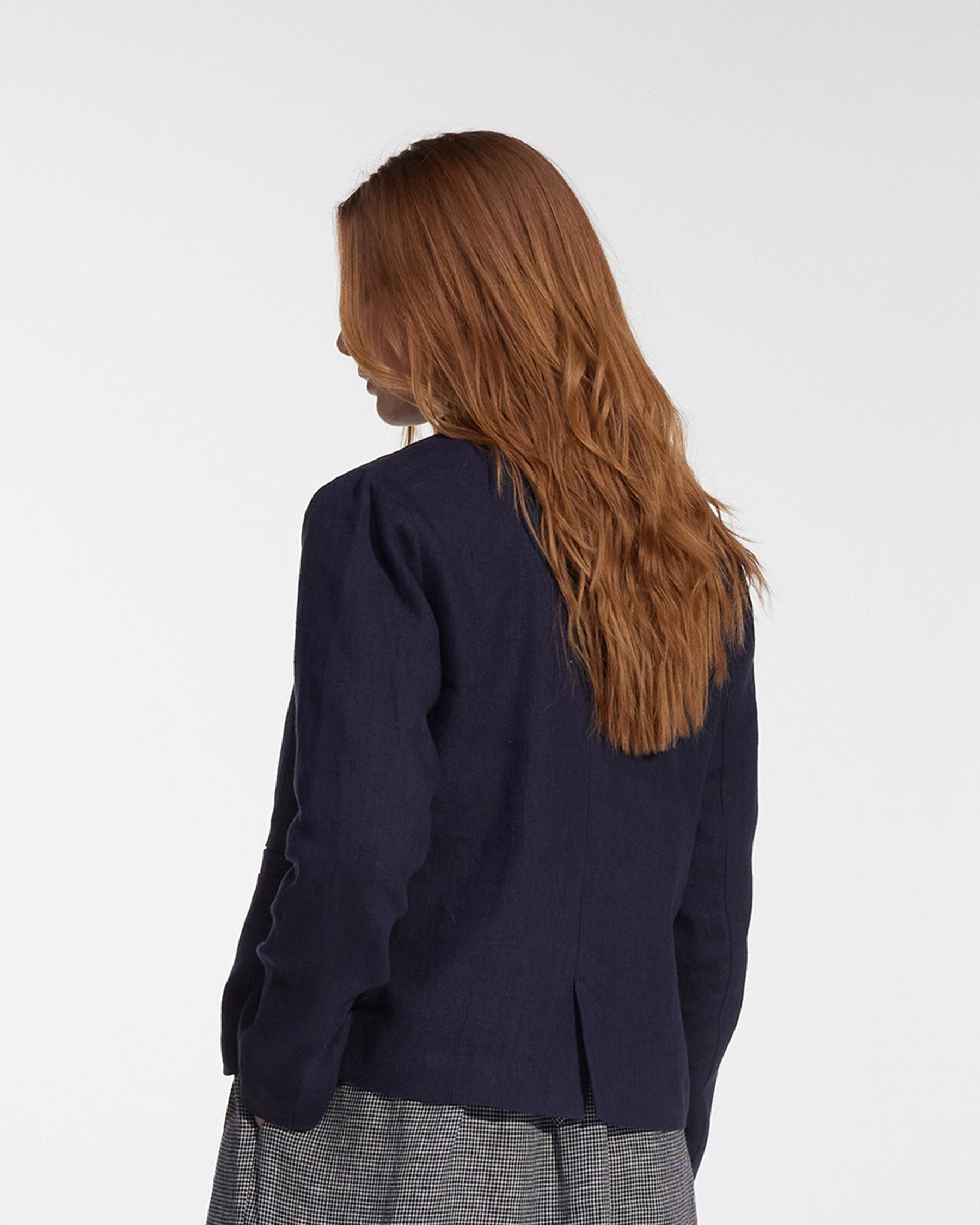 A person with long, wavy, light brown hair faces away from the camera, wearing the Pyne and Smith Clothiers Tailored Linen Jacket No.10 in Mariner Blue and a gray plaid skirt against a plain white background.